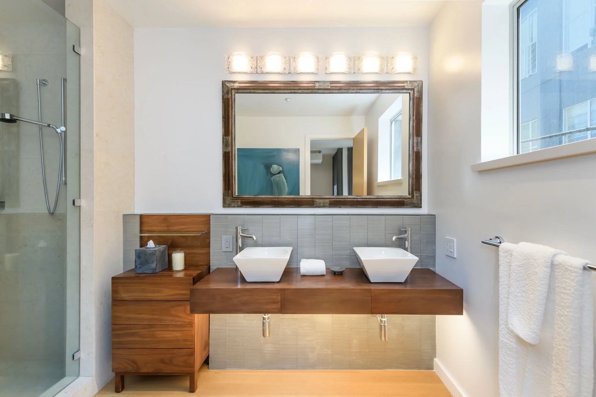 Modern bathroom with double white vessel sinks on a wooden vanity, large mirror with wooden frame, glass-enclosed shower on the left, window on the right, white walls, and bathroom accessories such as candles, tissue box, and towel.