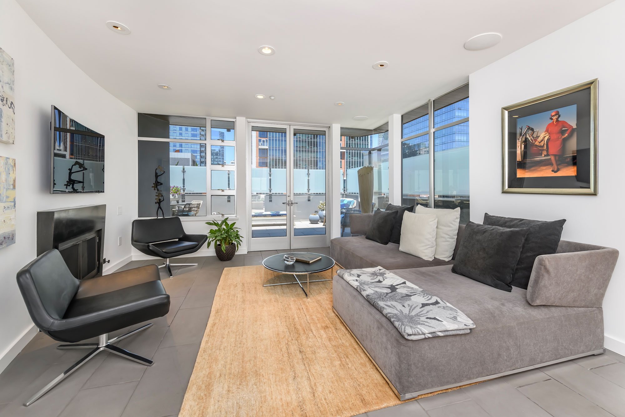 Bright living room with large glass windows, gray sofa, black chairs, small coffee table, potted plant, and wall art.