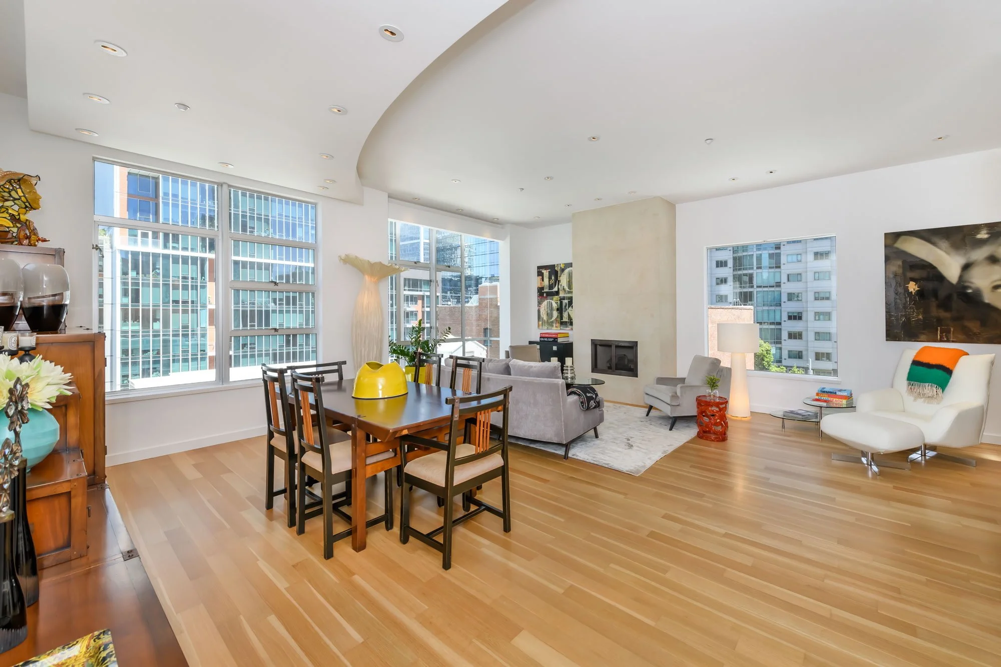 Modern living room with large windows, a gray sofa, white armchair, wooden dining table with black chairs, colorful vases, artwork, and cityscape view.