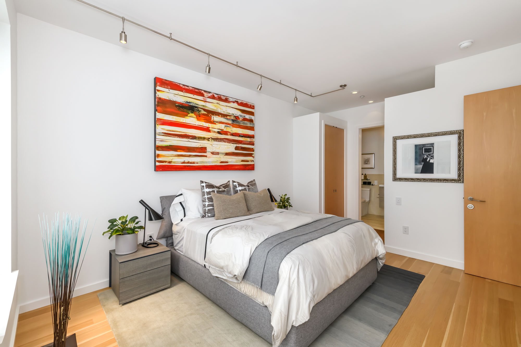 A modern bedroom with a bed, nightstand, and artwork on the walls. The bed has white and gray bedding with decorative pillows. There is a small side table with a plant and a lamp, and colorful abstract artwork hangs above the bed. A doorway leads to 