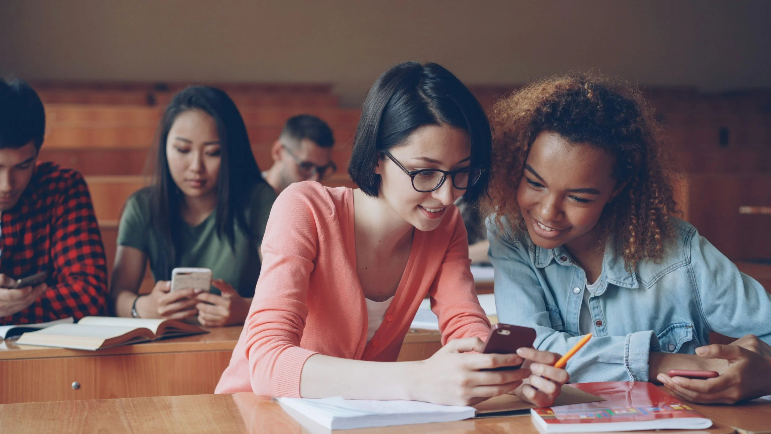 A diverse group of students sitting at a wooden desk in a classroom, looking at their phones and smiling.
