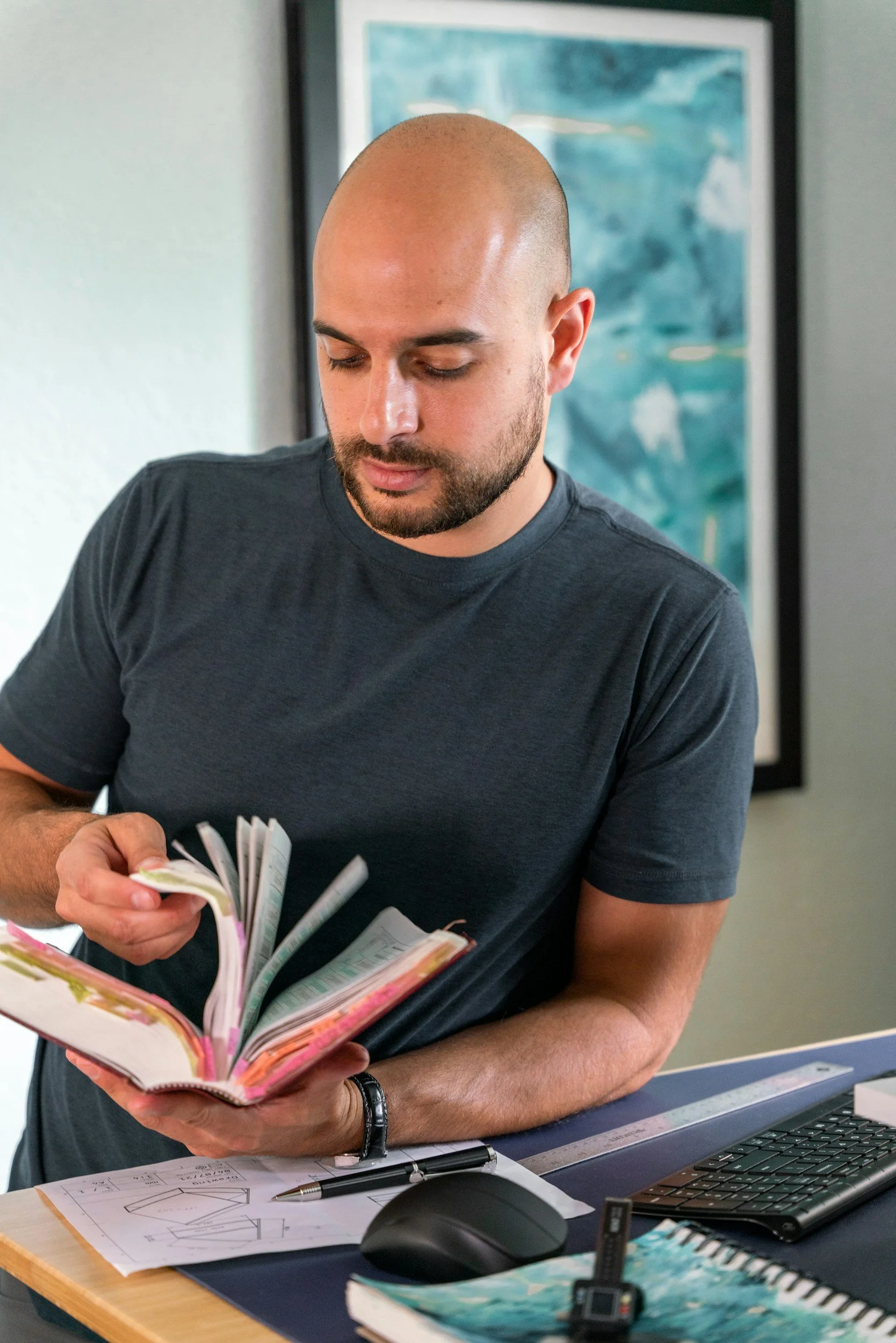 A man with a shaved head and beard looking down at a pink planner or notebook, holding it with both hands. The desk has papers, a pen, a computer mouse, a keyboard, and a small electronic device. In the background, there is a colorful abstract painting on the wall.