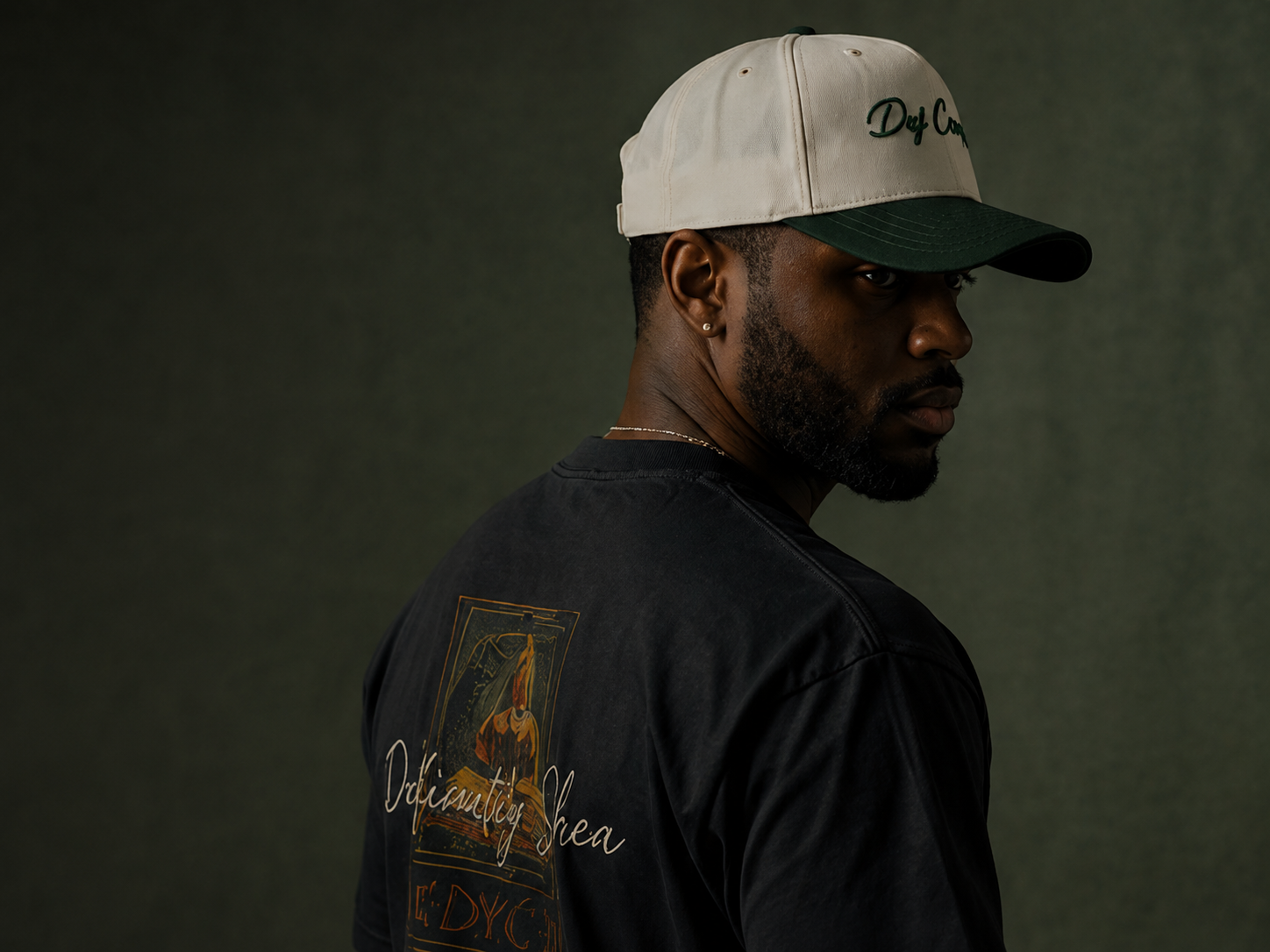 A man with dark skin, short hair, and a beard, wearing a beige and green cap, a black t-shirt with a graphic and text, and a small earring, looking over his shoulder against a dark green background.