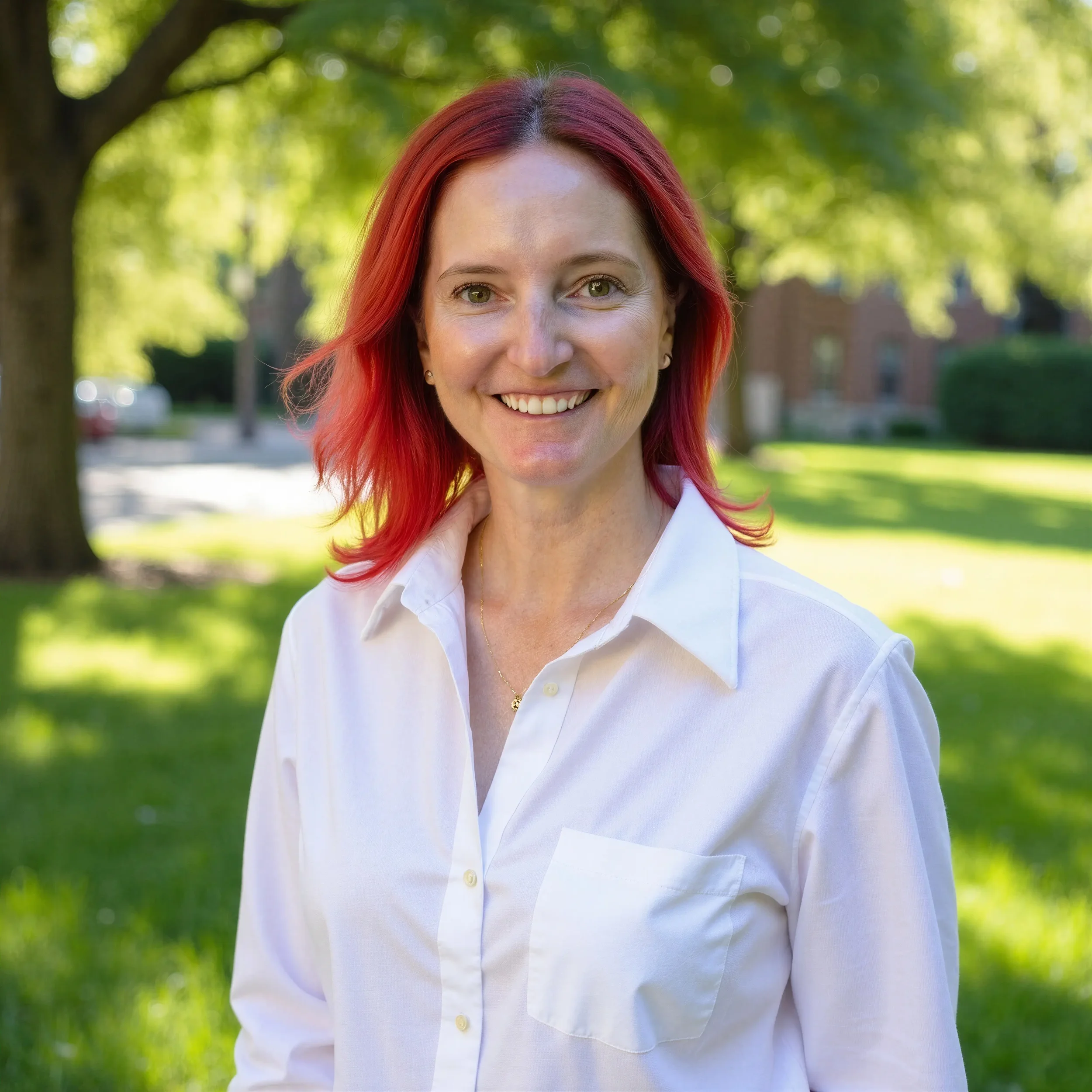 Close-up of a smiling woman with red hair, wearing a white shirt, standing outdoors with blurred green trees and grass in the background.