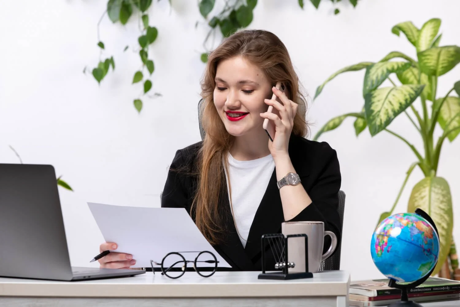 A woman sitting at a desk, talking on her cellphone, and looking at a paper, with a laptop, a globe, and books on the desk, and plants in the background.