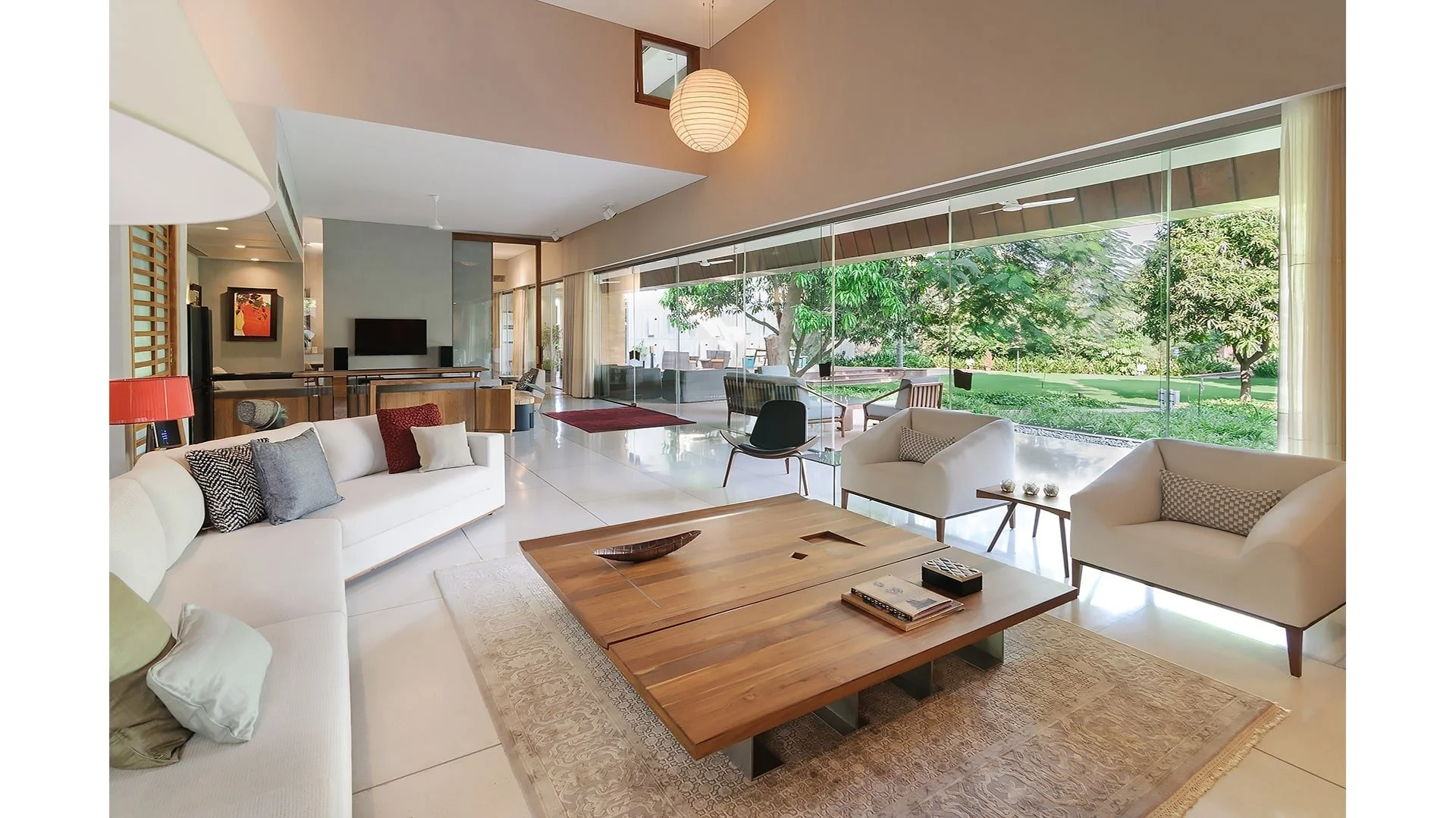 Interior of a modern living room with white sofa, variety of pillows, wooden coffee table, and view of lush green outdoor area through large glass windows.