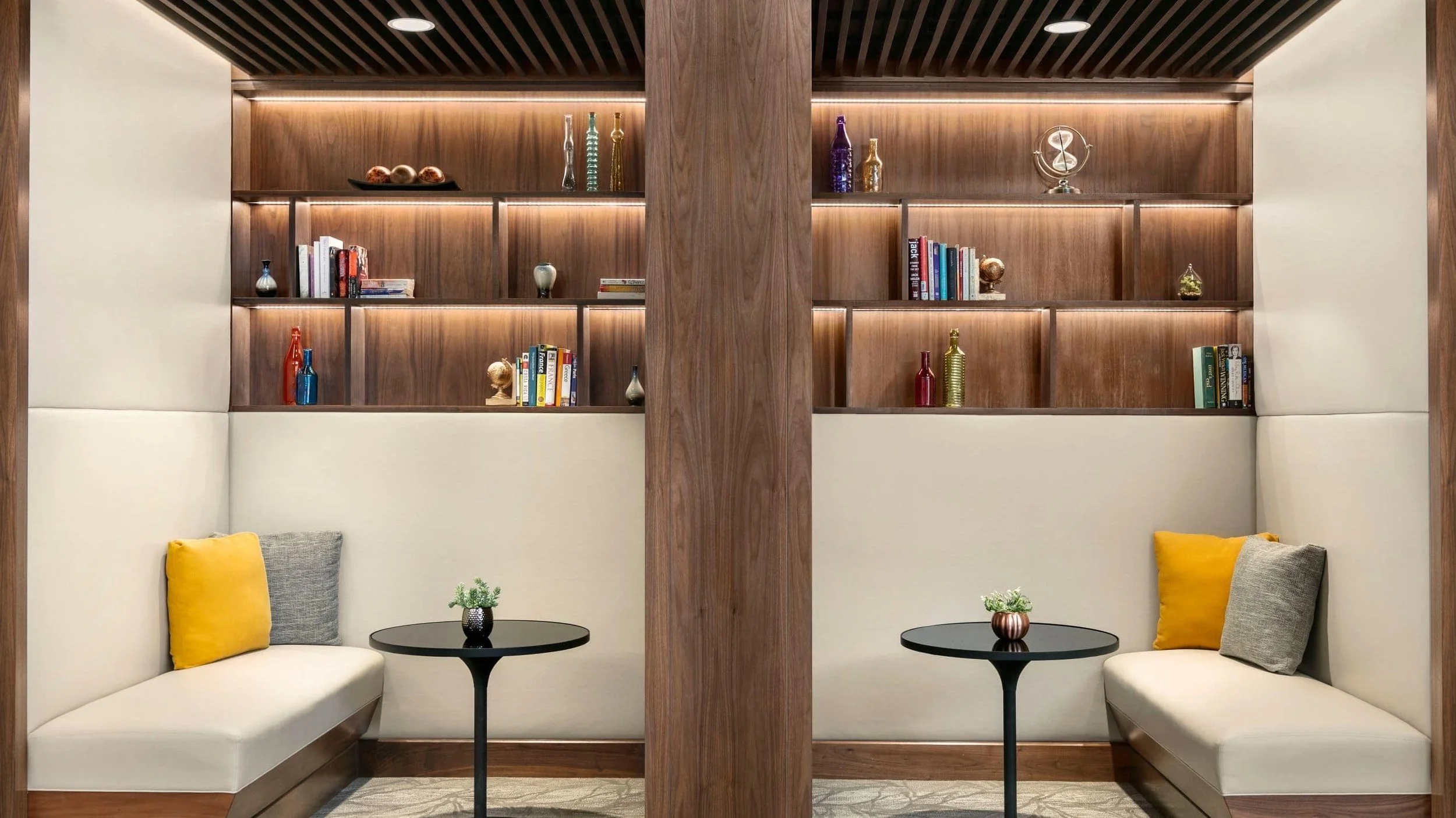 A cozy seating area with white upholstered bench seating, yellow and gray pillows, black round tables, and a wooden bookshelf with decorative bottles, books, and art pieces behind.