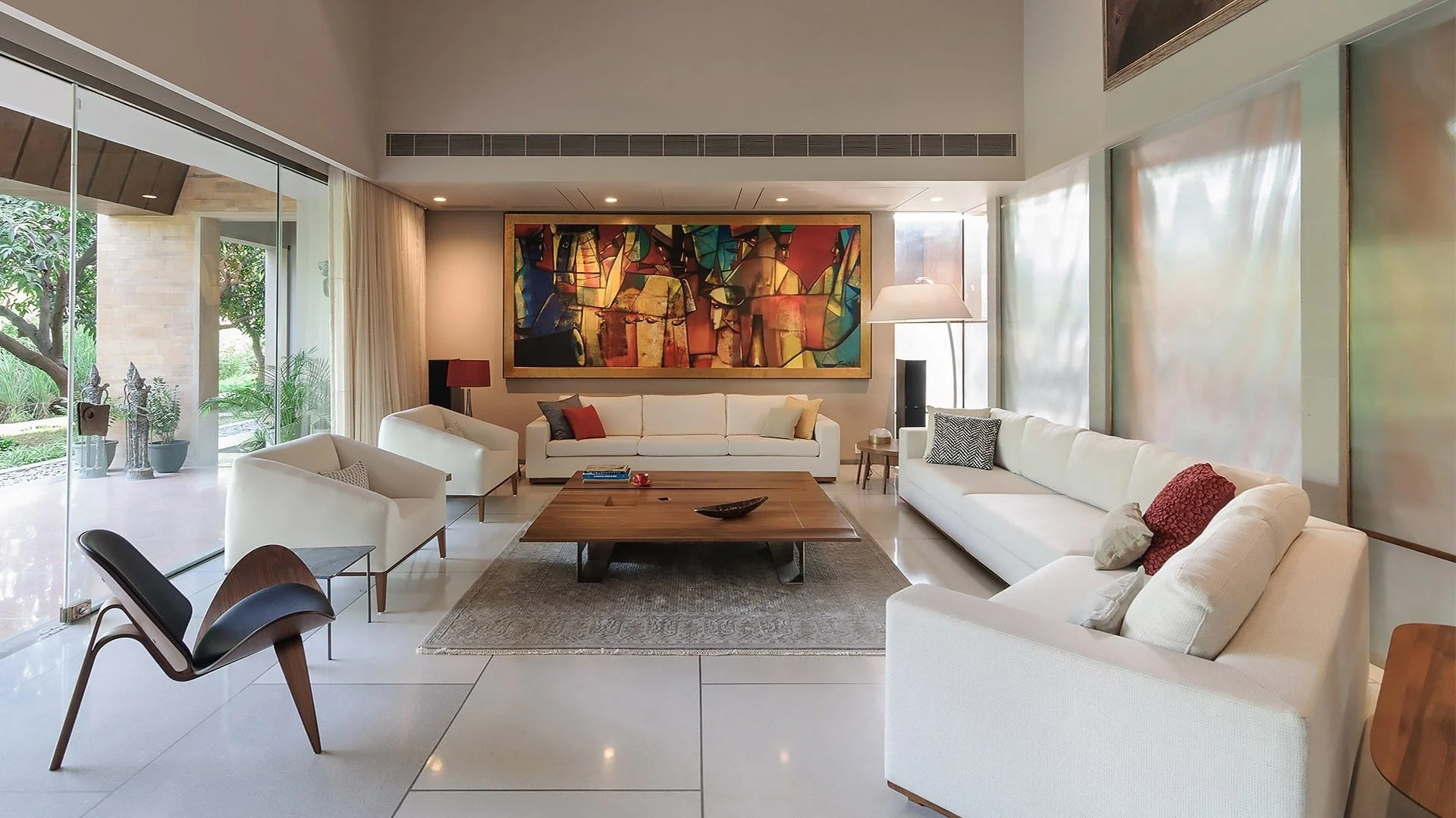 Modern living room with white sofas, a wooden coffee table, colorful abstract art, and large windows with sheer curtains and outdoor greenery