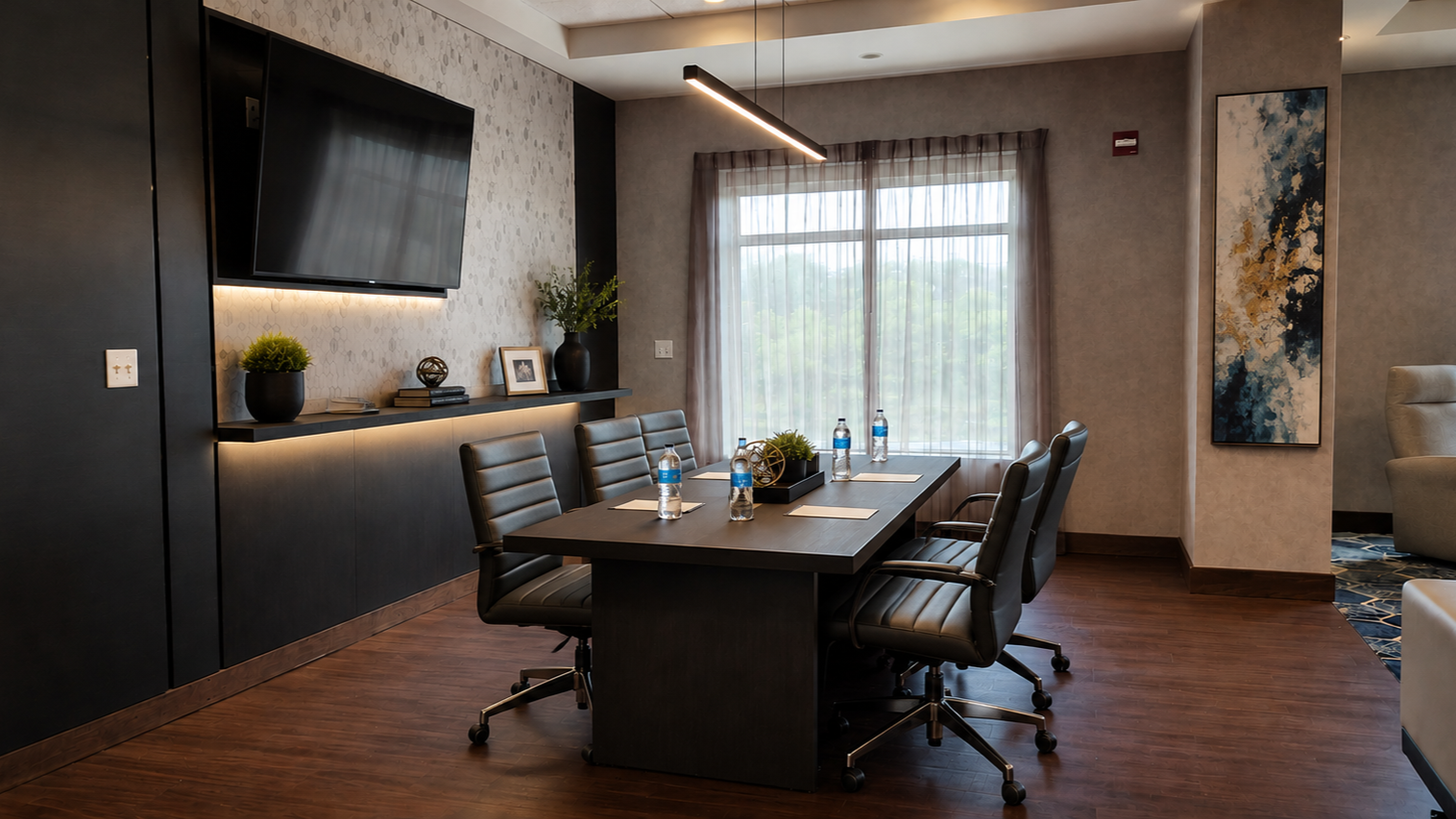 Conference room with a table and six chairs, a wall-mounted TV, a window with sheer curtains, decorative plants, and abstract artwork.