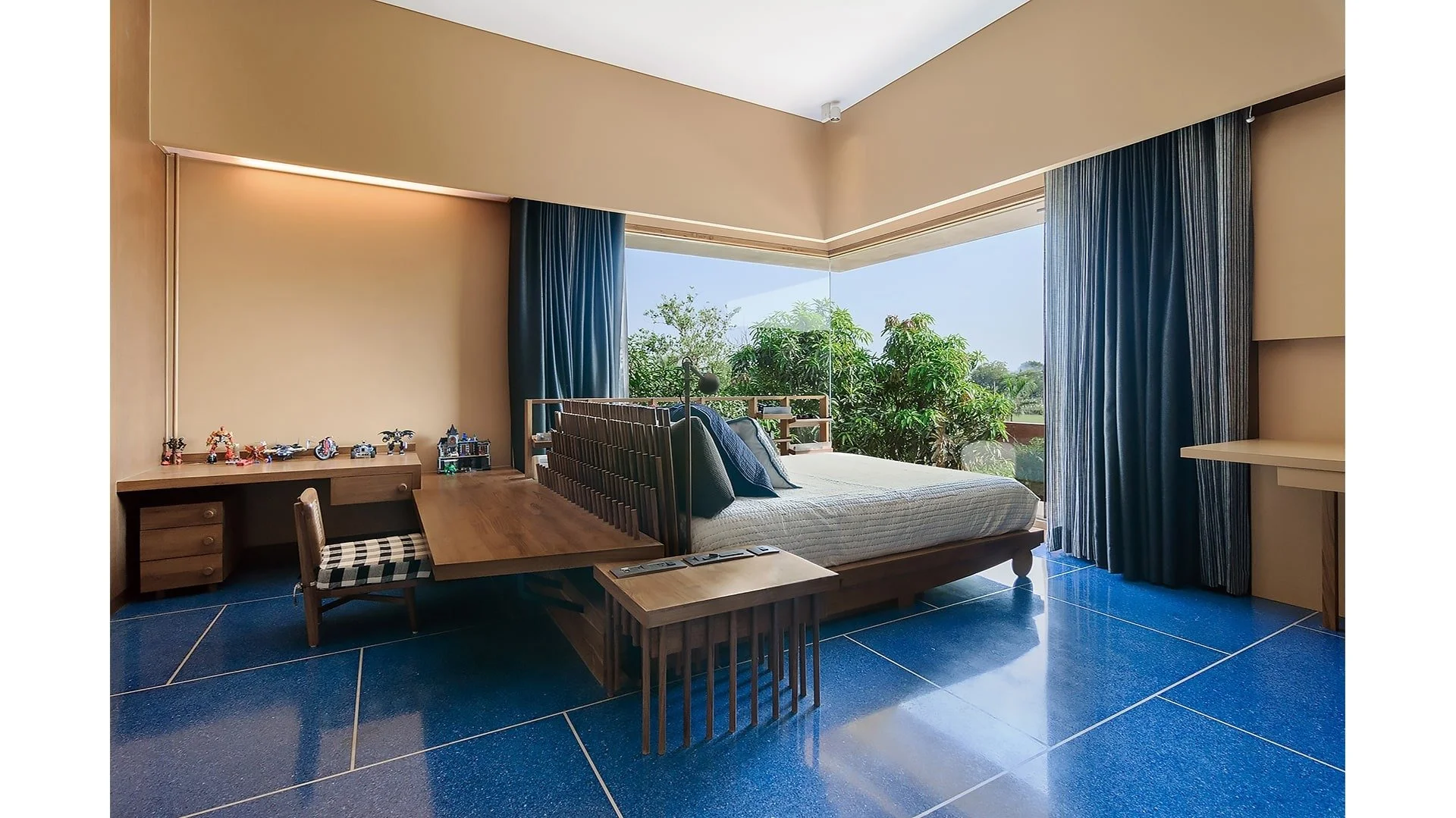 A bedroom with a bed near a large window with blue curtains, overlooking trees outside. There is a wooden desk with toys and a chair, and a small table at the foot of the bed. The floor is tiled in dark blue.