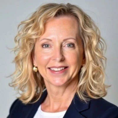 Portrait of a woman with curly blonde hair, wearing a navy blazer and white top, smiling against a gray background.
