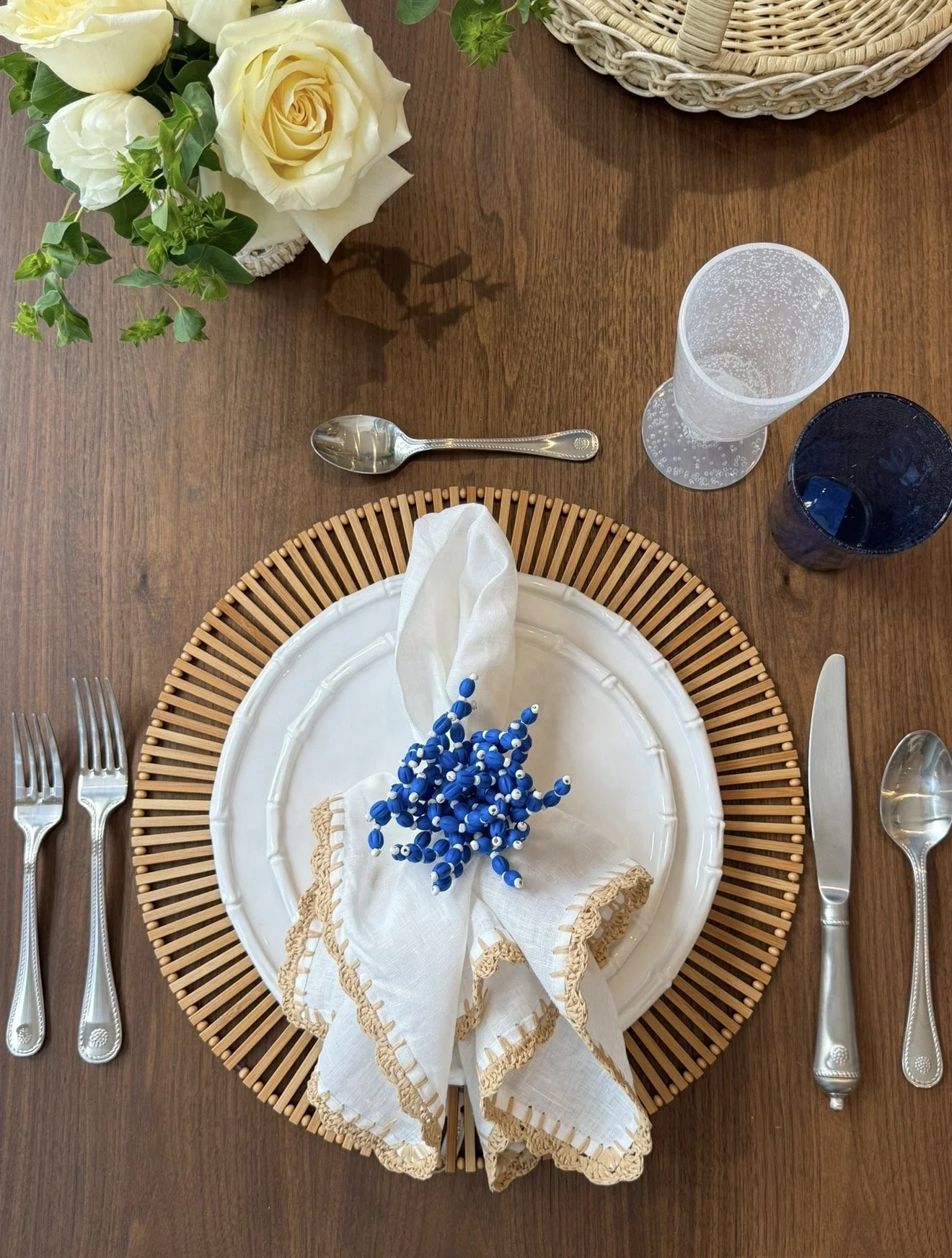 Palm Beach. Table setting with white plate, decorative beige charger, napkin with blue and white beads, silverware, and a floral centerpiece with white and green flowers, on a wooden table.
