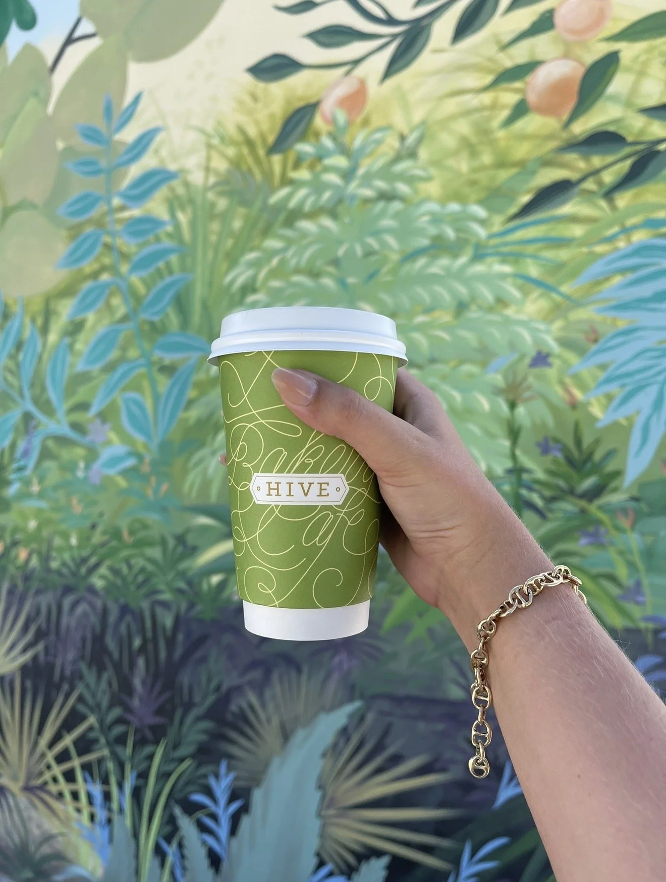 Palm Beach. A person with a gold bracelet on their wrist holds a green paper coffee cup labeled 'HIVE' in front of a botanical mural featuring large green leaves and plants.