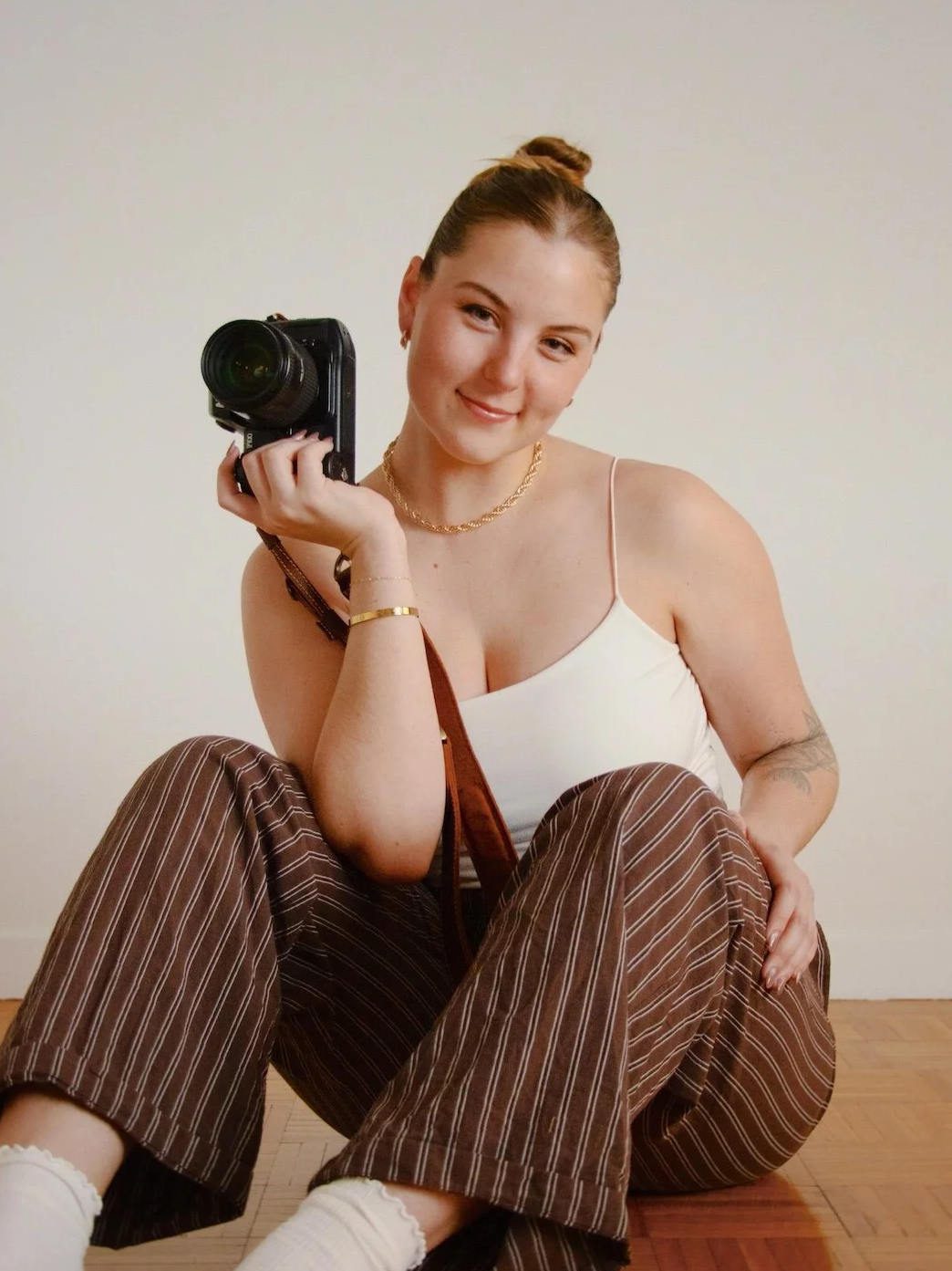 Social Media Strategist & Marketing Manager, Ingrid Marinak. A young woman with dark blonde hair in a bun, smiling, holding a camera. She wears a white tank top, striped brown pants, and white socks, sitting on a wooden floor against a plain wall.