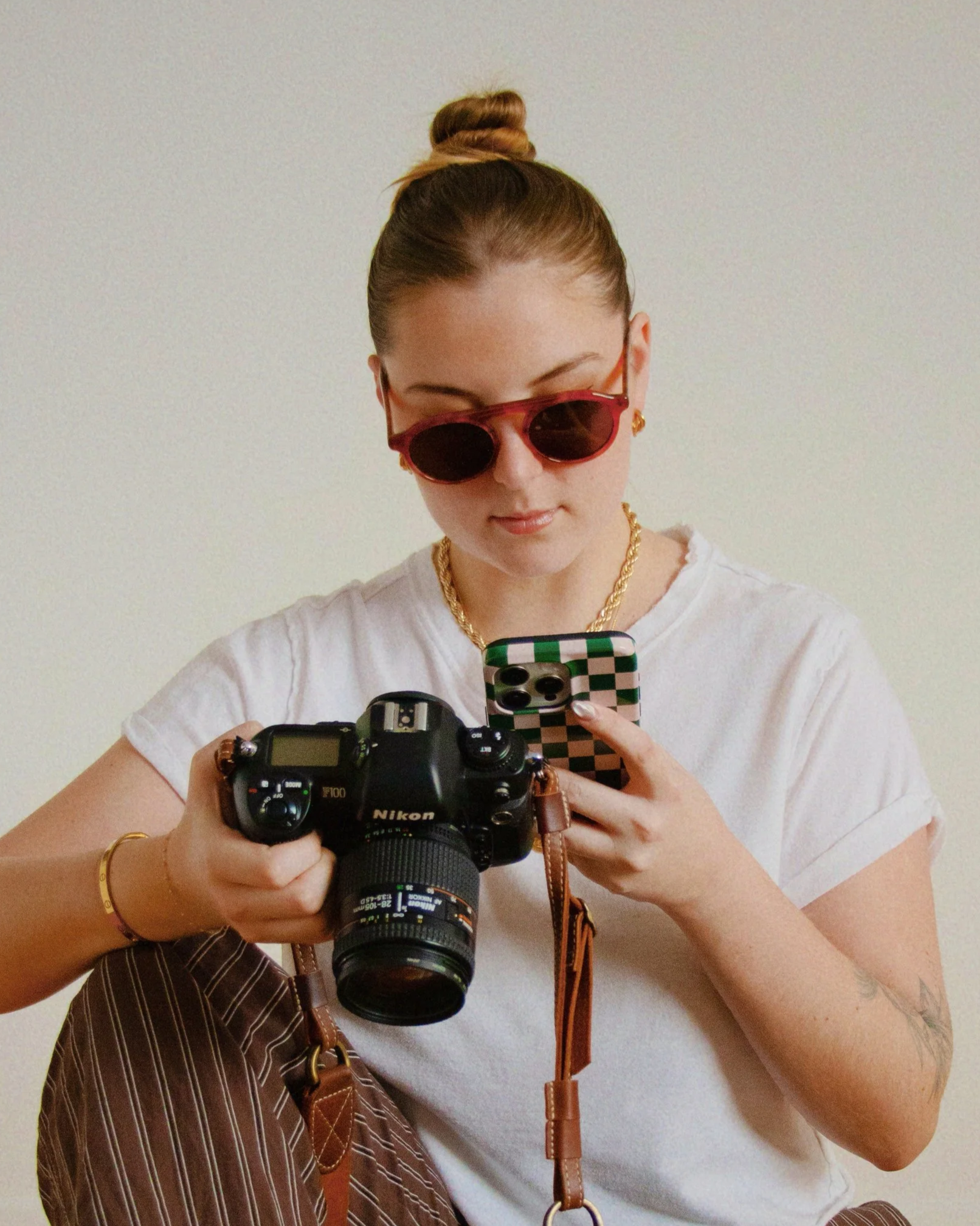 Ingrid Marinak, Social Media Strategist & Marketer. Young woman working on her iPhone as well as holding a professional camera, wearing red sunglasses, a white t-shirt, gold jewelry against a plain wall.