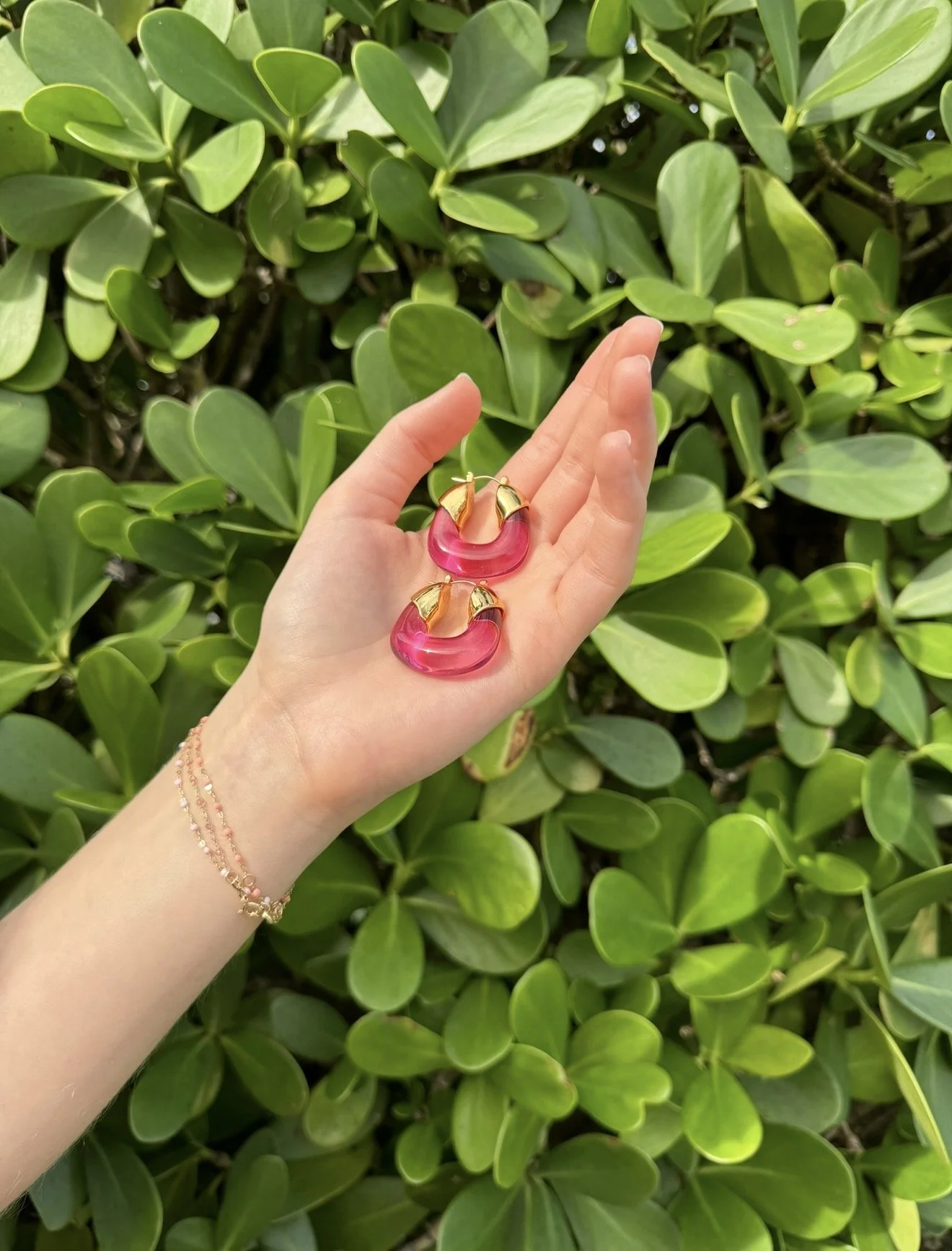 Palm Beach. A person's hand holding pink and gold earrings in front of green leafy bushes.