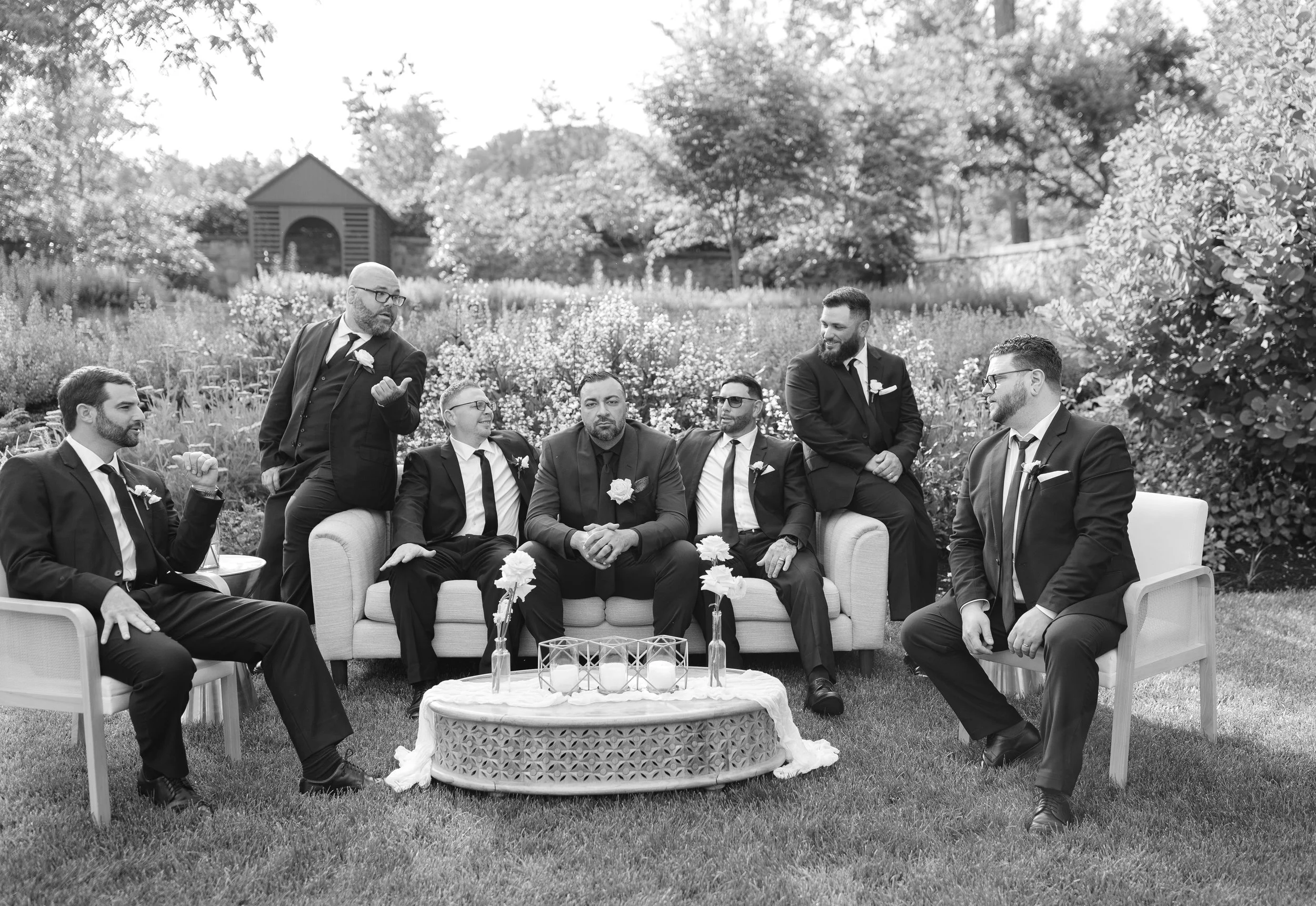 Black and white photo of a group of men in suits at a wedding outdoor ceremony, sitting and standing around a sofa and coffee table with candles, in a garden setting with trees and flowers in the background.