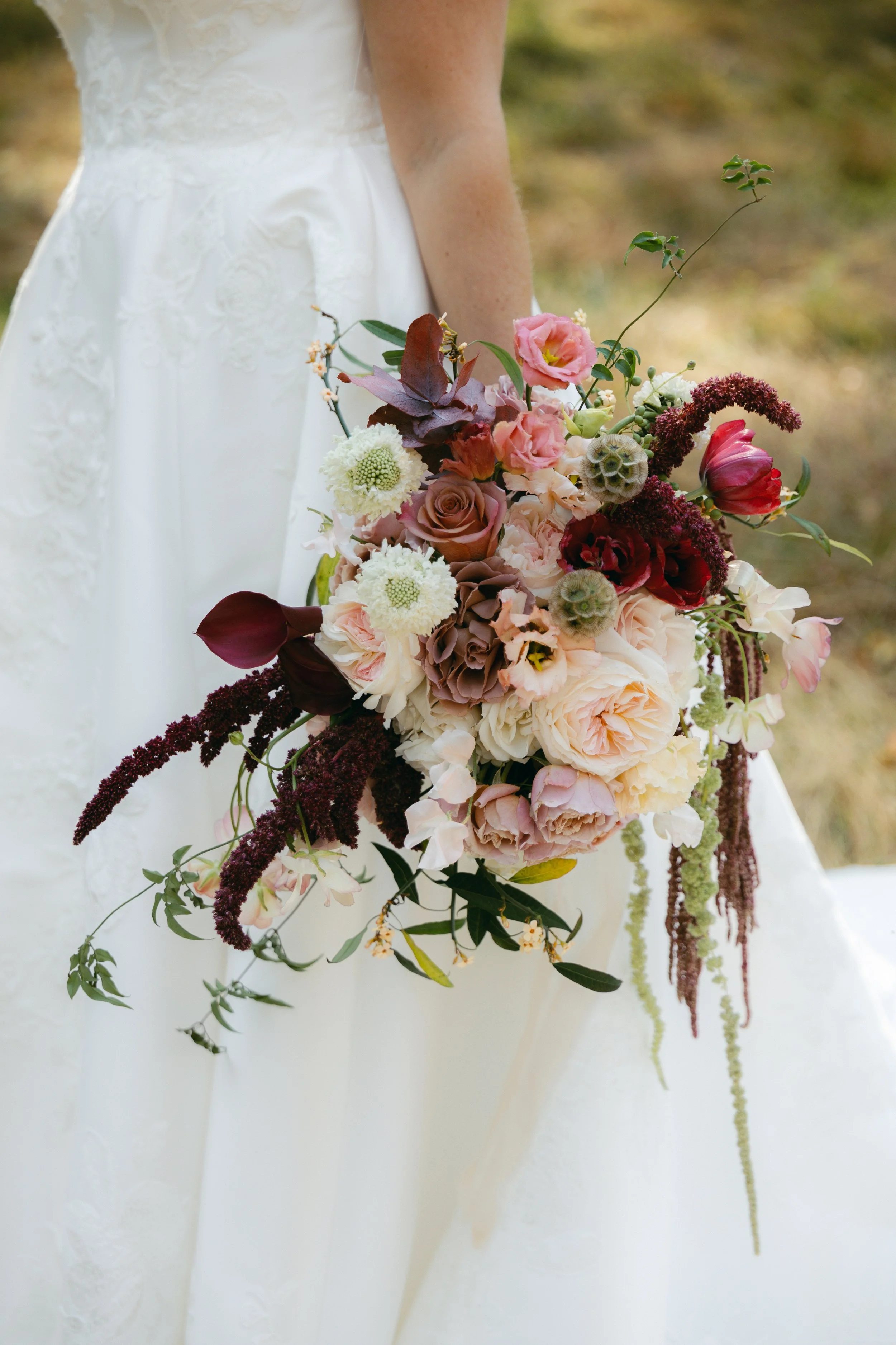 A bride holding a large bouquet of various flowers, standing outdoors. The bride's white wedding dress is visible from the waist down.