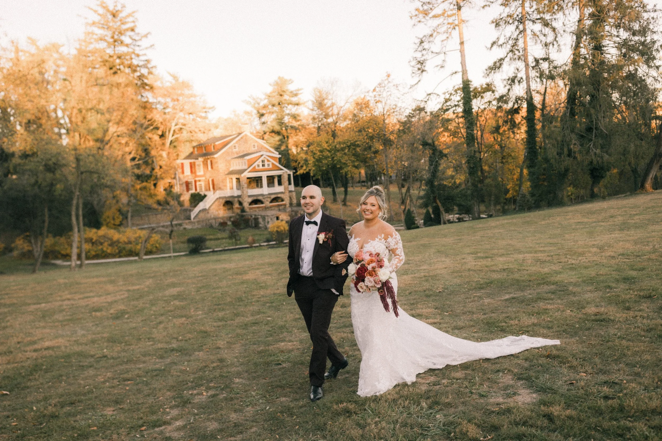 A bride and groom walking together on a grassy area during sunset, with trees and a large house in the background. The bride is wearing a white wedding gown and carrying a bouquet of flowers, while the groom is dressed in a black tuxedo.
