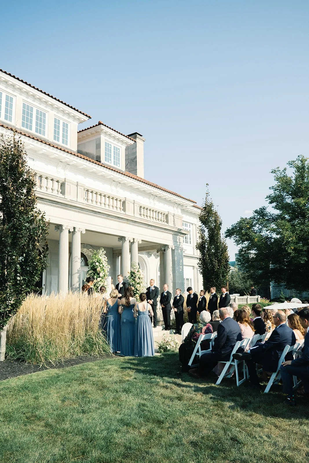 Wedding ceremony taking place outdoors in front of a large, white mansion with tall columns, with guests seated on white chairs observing the event.
