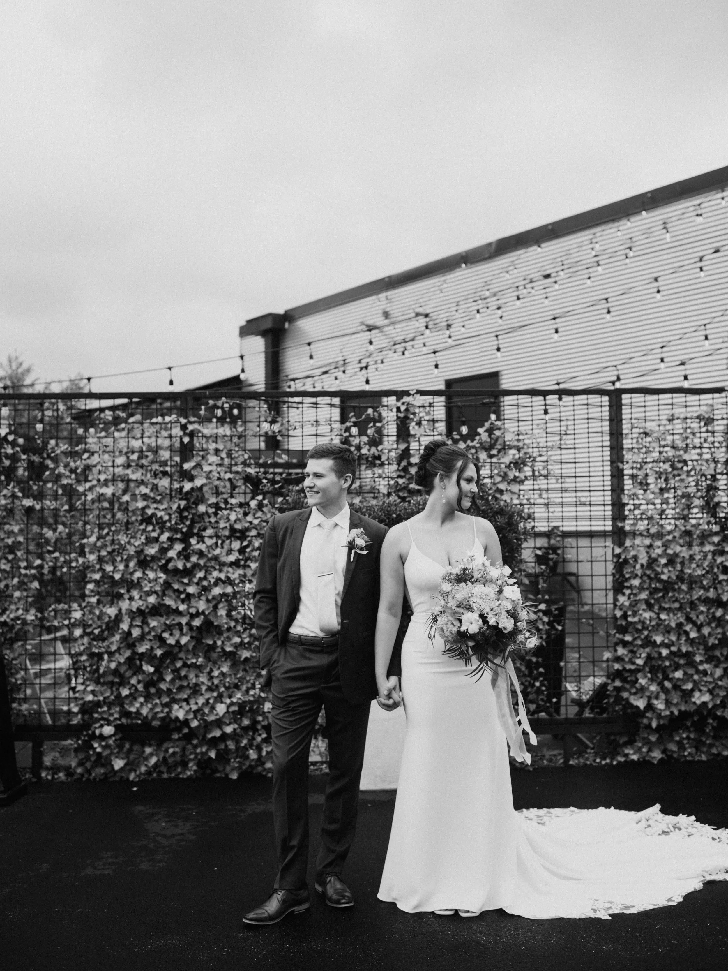 A black and white photo of a bride and groom holding hands, walking outdoors. The bride wears a white wedding dress and holds a bouquet. The groom wears a dark suit with a boutonniere. They are standing in front of a fence with foliage and string lights, and a building with horizontal siding is visible in the background.