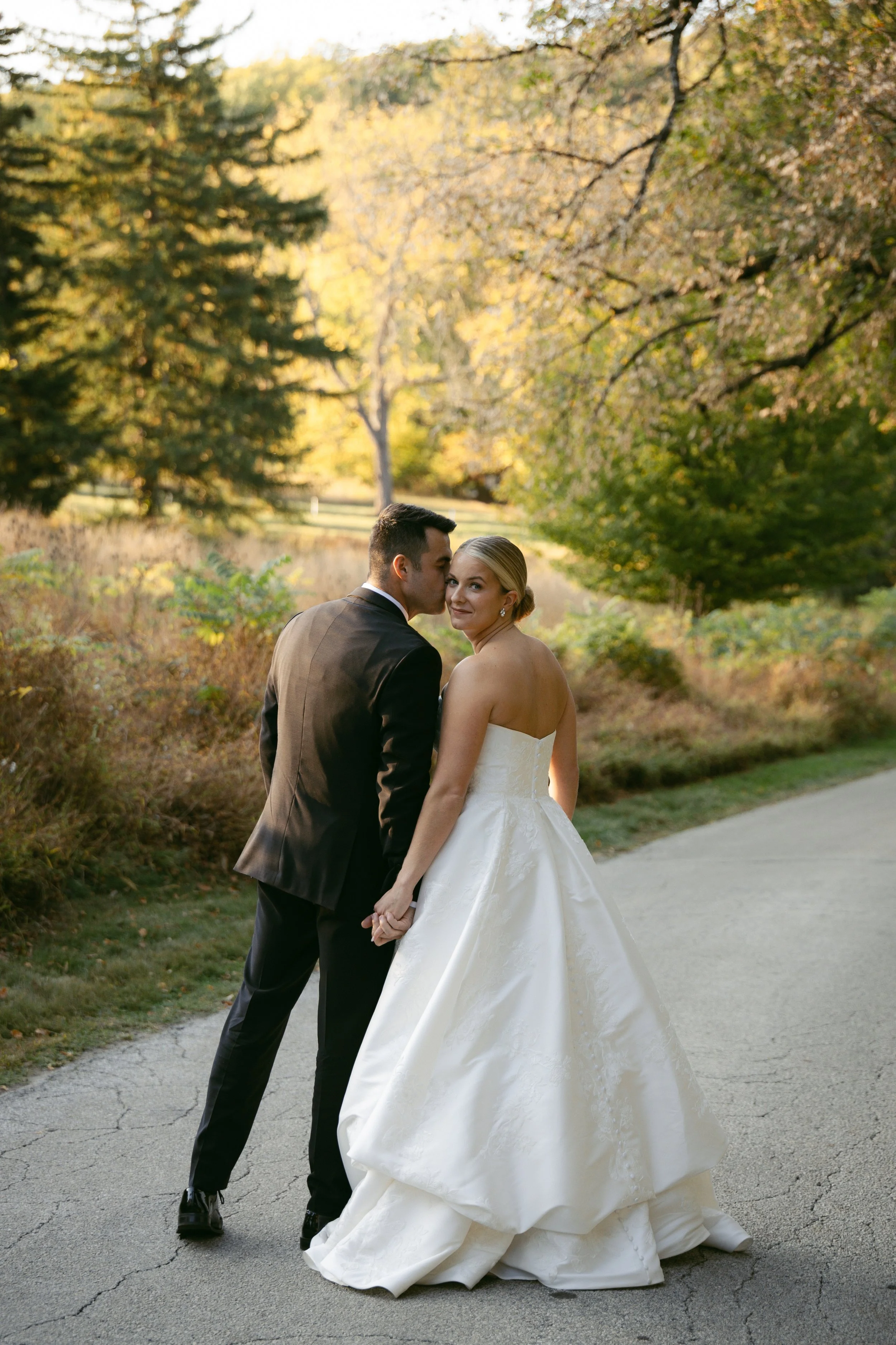 A newlywed couple holding hands on a paved path in a park with autumn foliage. The groom is dressed in a black suit and the bride is in a strapless white wedding gown, looking at the camera.