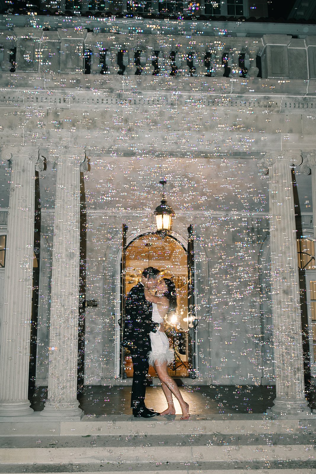A couple dressed in formal attire sharing an intimate moment in front of a building with classical columns at night. The photo is covered with sparkling light effects, creating a romantic atmosphere.