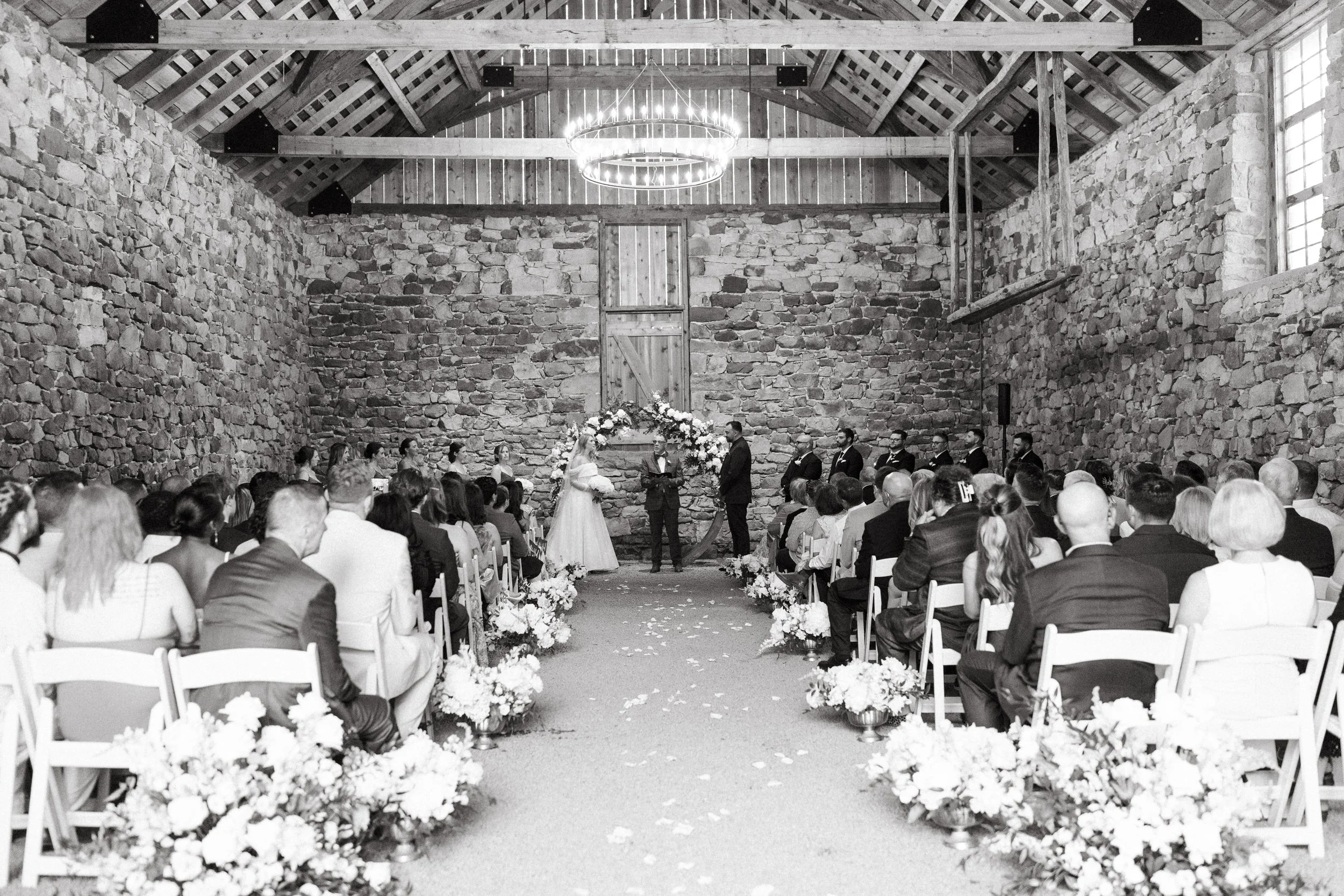 A black-and-white photo of a wedding ceremony inside a rustic barn with stone walls and a wooden ceiling. The bride and groom stand facing each other in front of an officiant, under a floral arch. Guests are seated on white chairs along an aisle decorated with white flowers.