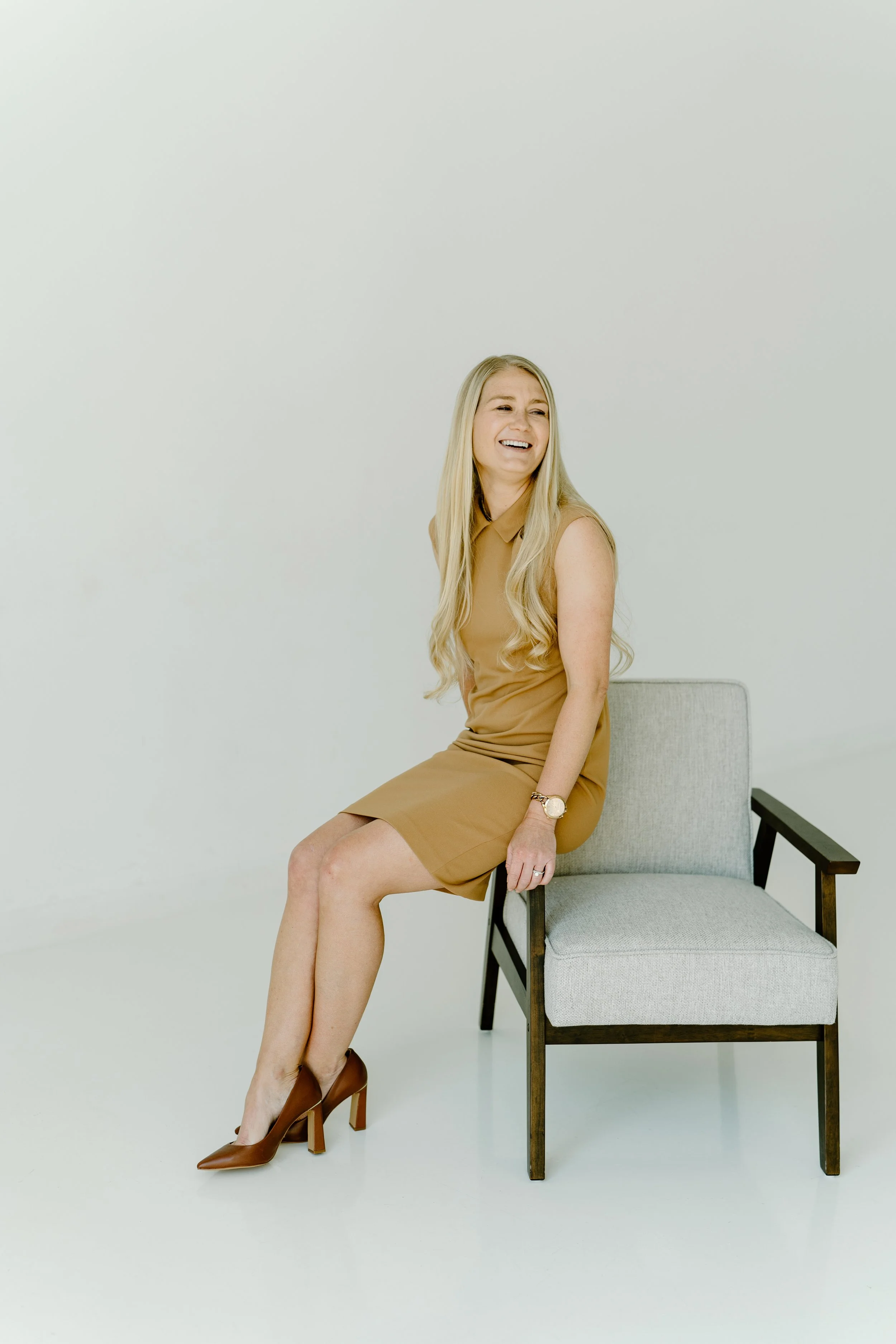 A woman with long blonde hair sitting on a gray chair, wearing a tan sleeveless dress and high heels, smiling and looking to her right against a plain white background.