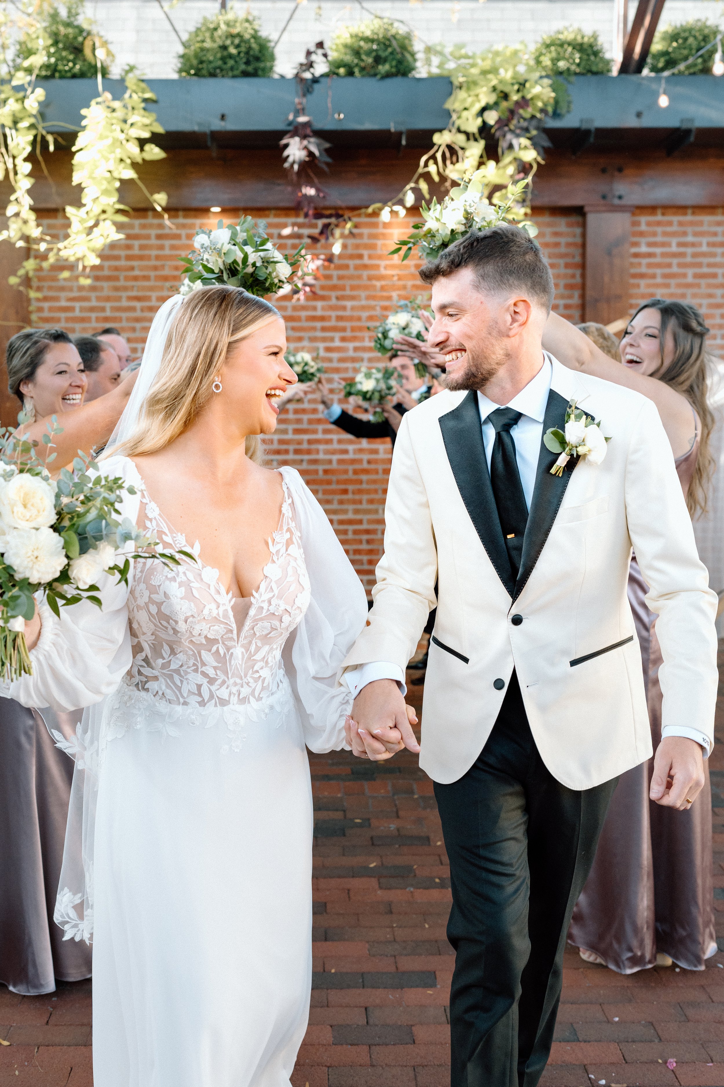 A bride and groom holding hands and smiling at each other during their wedding reception, surrounded by bridesmaids holding bouquets, in a decorated outdoor setting with string lights and greenery.
