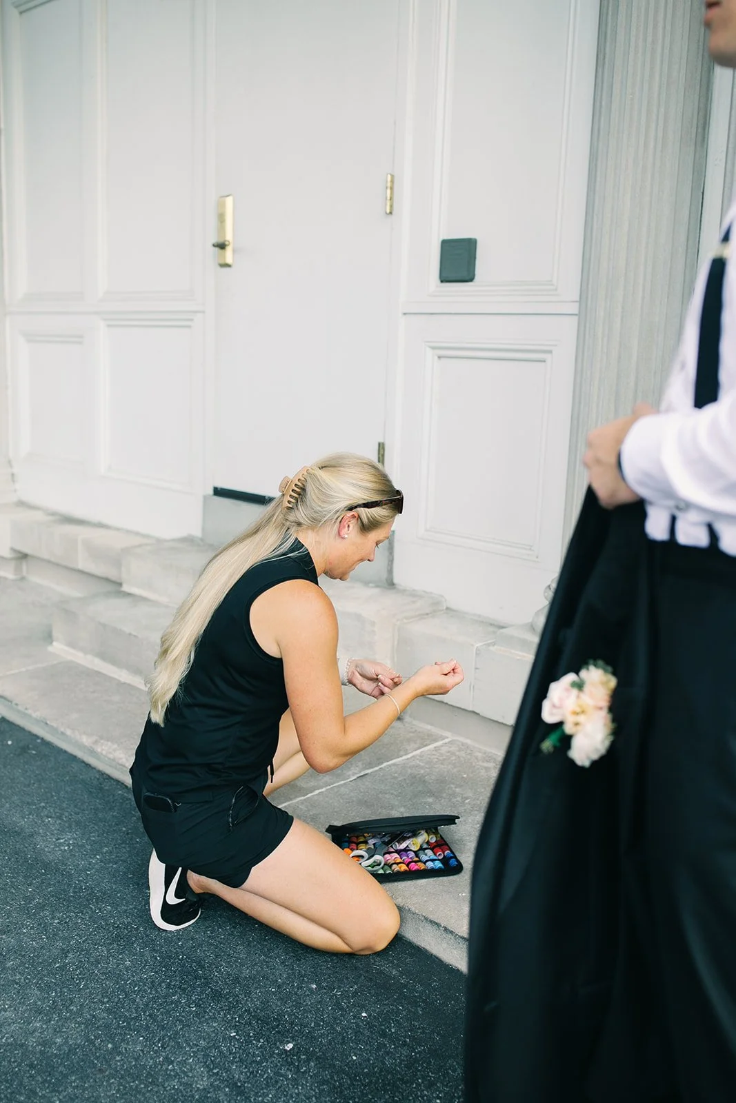 Kneeling woman with long blonde hair, wearing black shorts and a sleeveless top, organizing sewing or embroidery thread spools on the ground outside a white building.