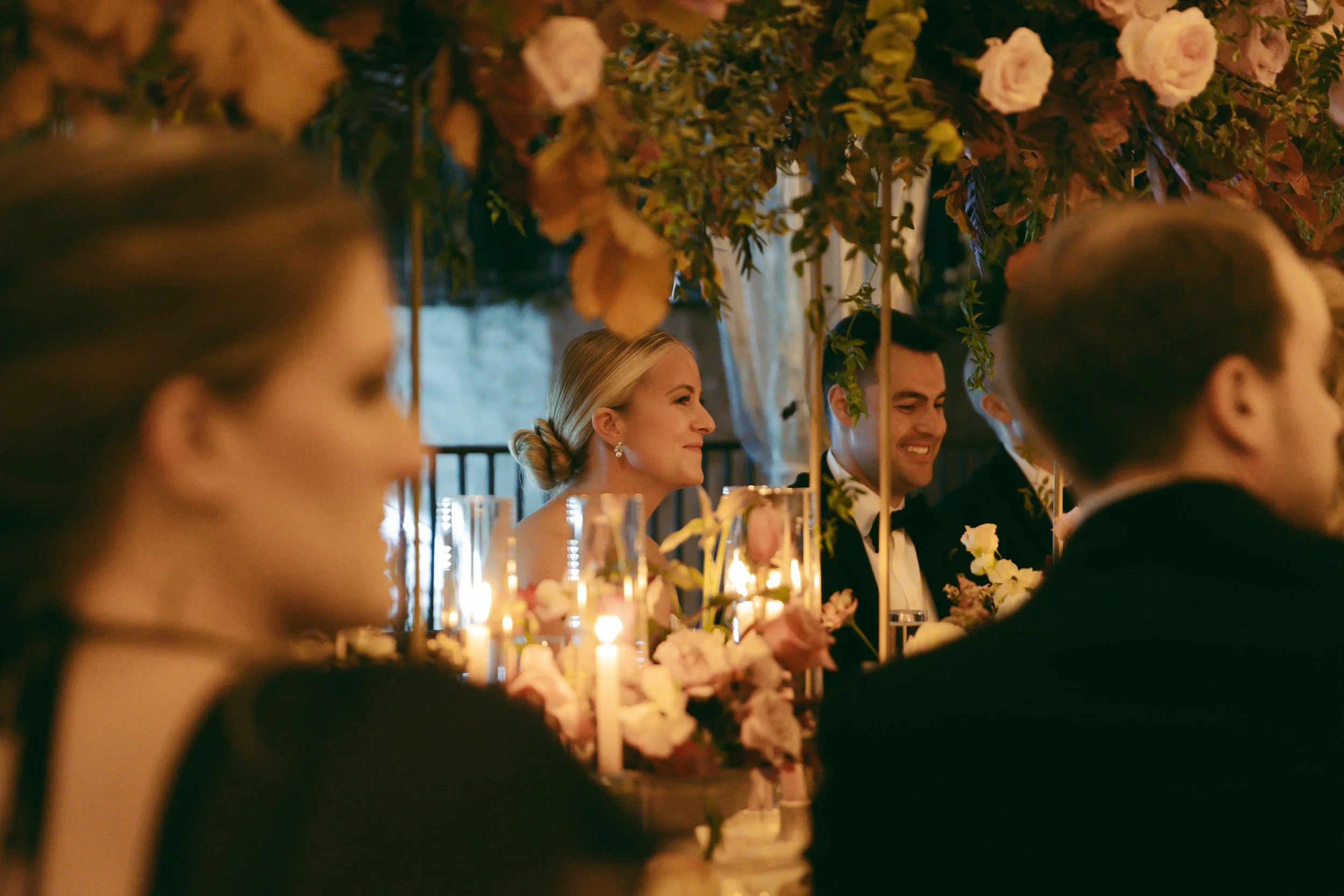 People sitting at a decorated wedding reception table, with floral arrangements and candles, smiling and engaged in conversation.