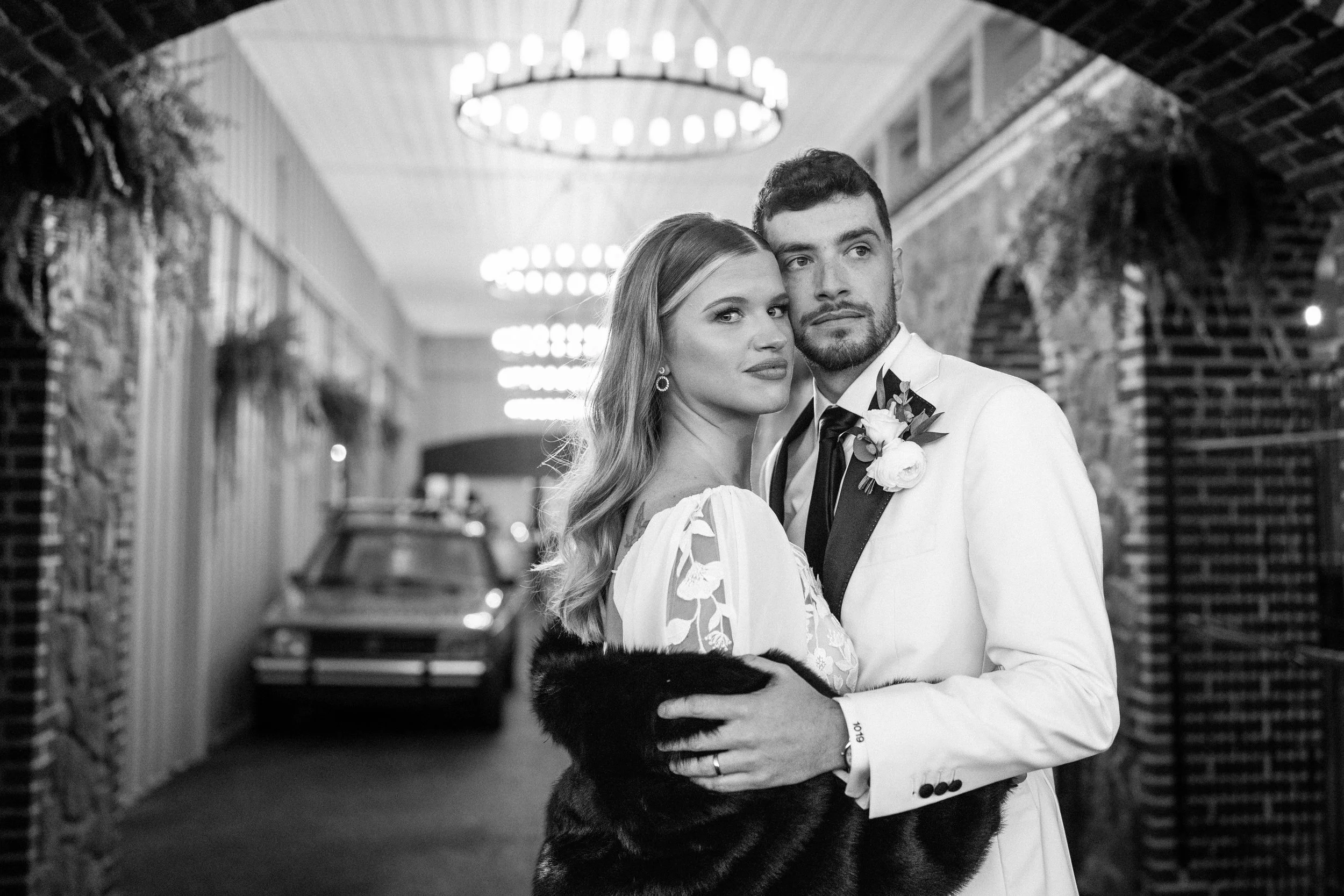 A black and white photo of a couple in formal wedding attire standing close together in an indoor setting with brick walls and decorative lighting.