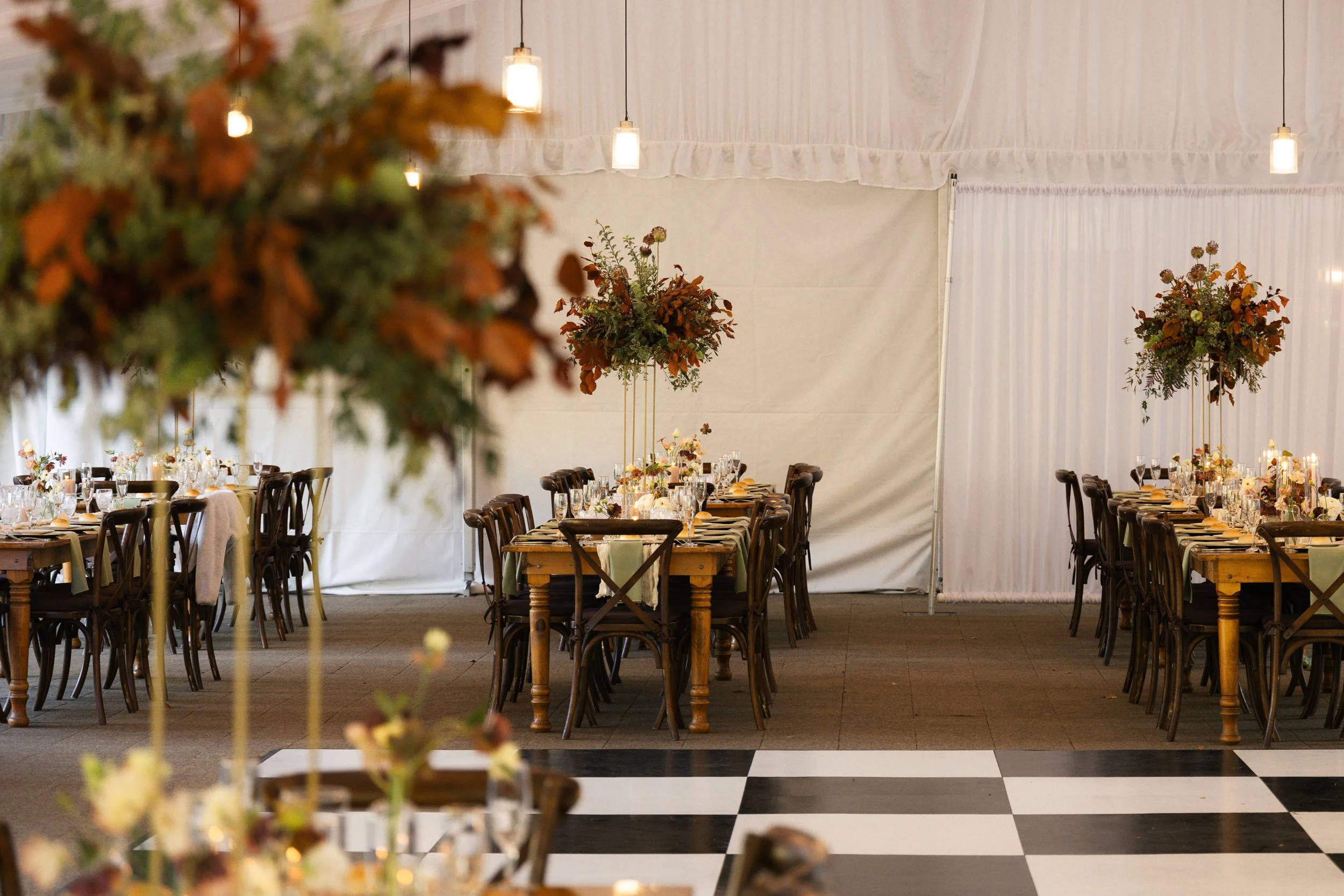 Wedding reception setup with long tables decorated with floral centerpieces, glassware, and candles in a large indoor venue with high ceilings, hanging lights, and a checkered black and white floor.