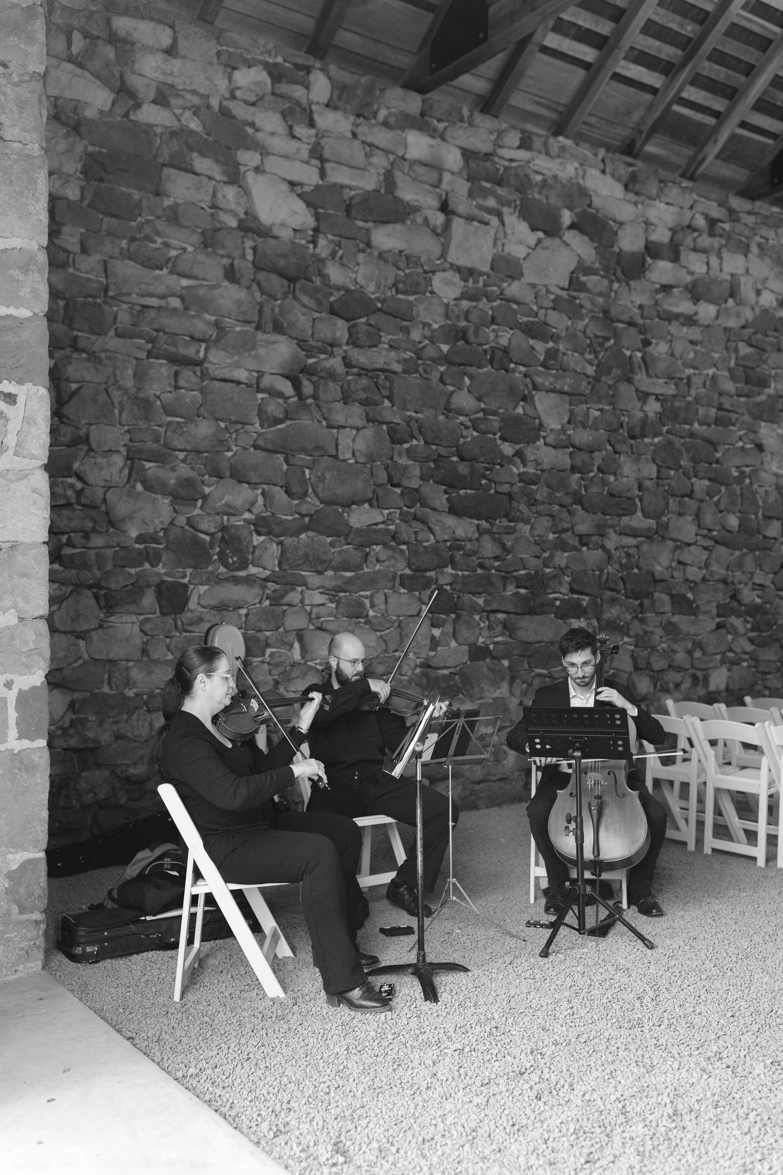 A group of three musicians playing string instruments in a rustic indoor setting with a stone wall in the background.