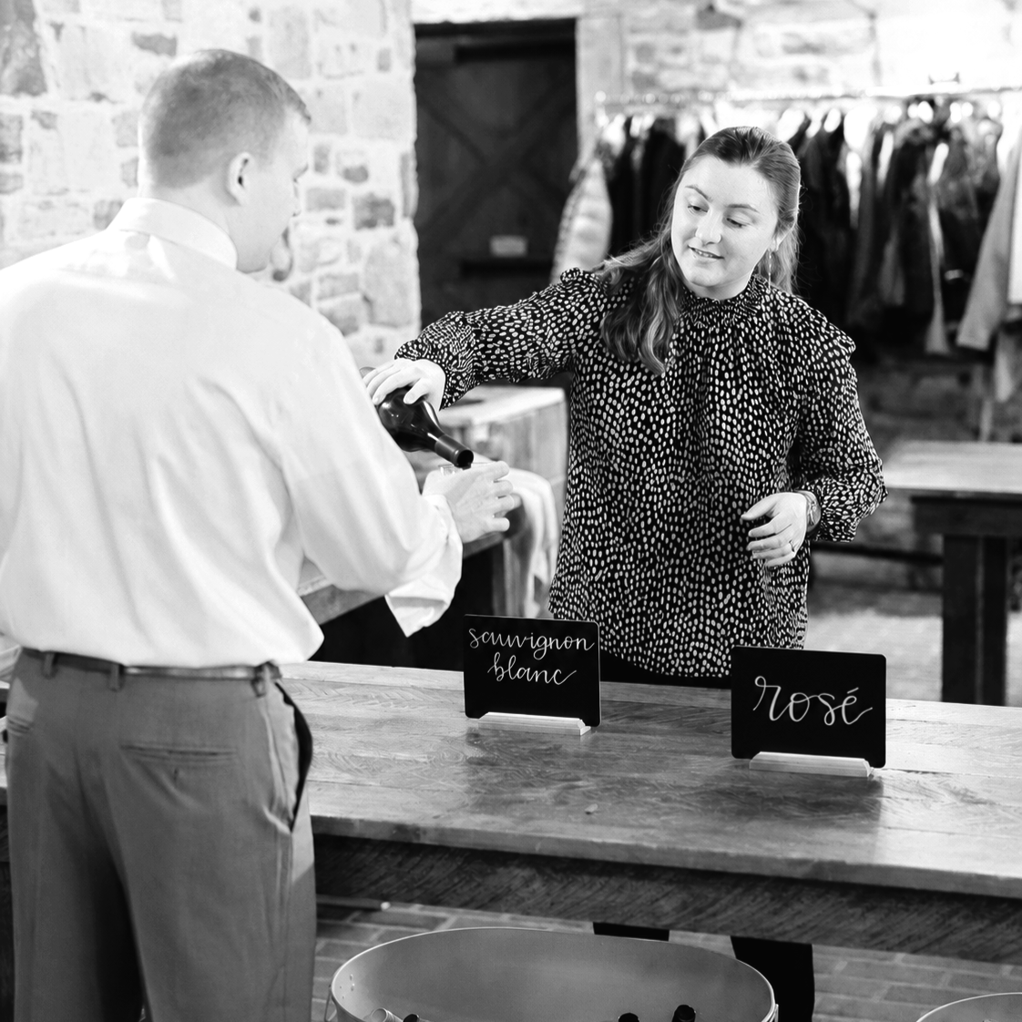 A woman is pouring wine into a glass held by a man at a wine tasting event. Signs on the table indicate different types of wine, such as sauvignon blanc and rosé.