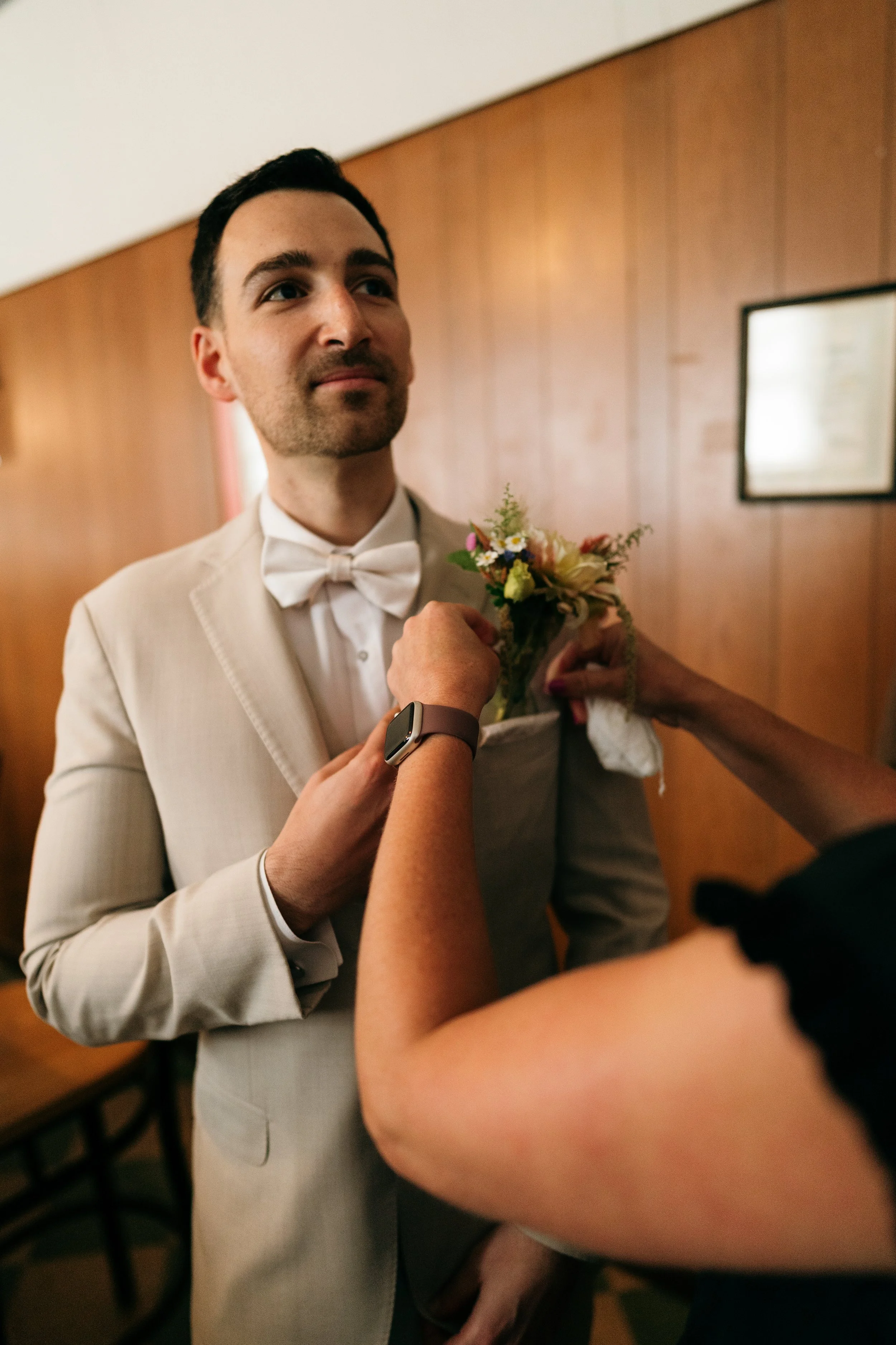 Groom in a beige suit and white bow tie adjusting his boutonniere, with close-up of a woman’s arm pinning it on, in an indoor setting with wooden wall panels.