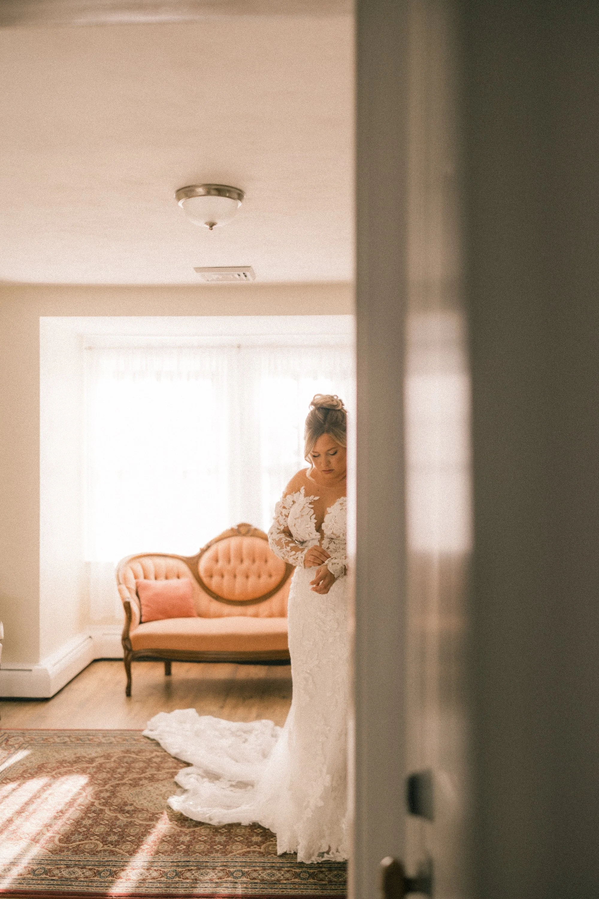 A bride in a lace wedding dress stands near a peach-colored vintage sofa in a warmly lit room with cream walls and patterned carpet.