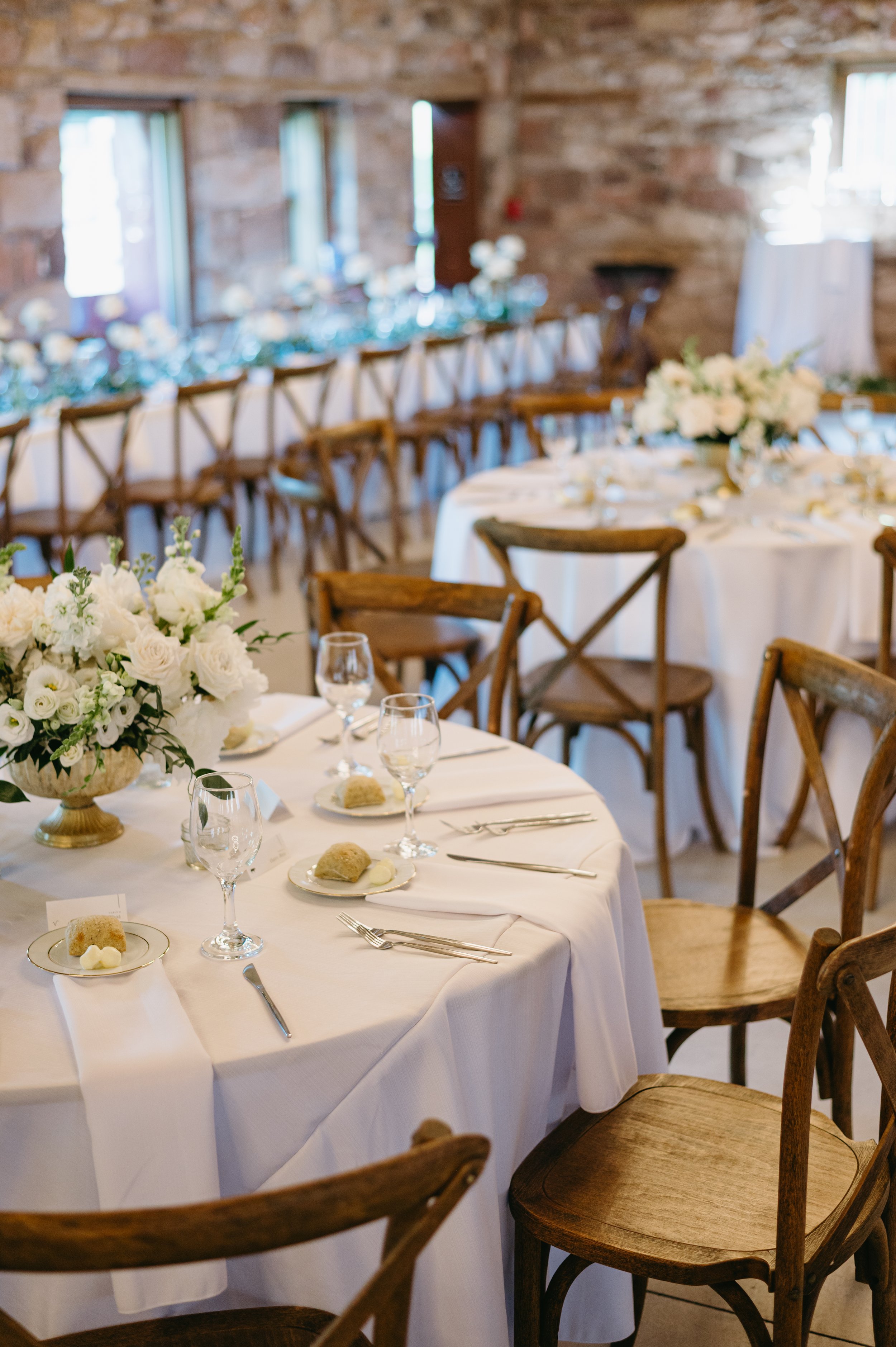A dining room with a long table decorated with white tablecloths, floral centerpieces, wine glasses, and plates with rolls. Wooden chairs are arranged around the table, and there are windows with natural light and brick walls in the background.