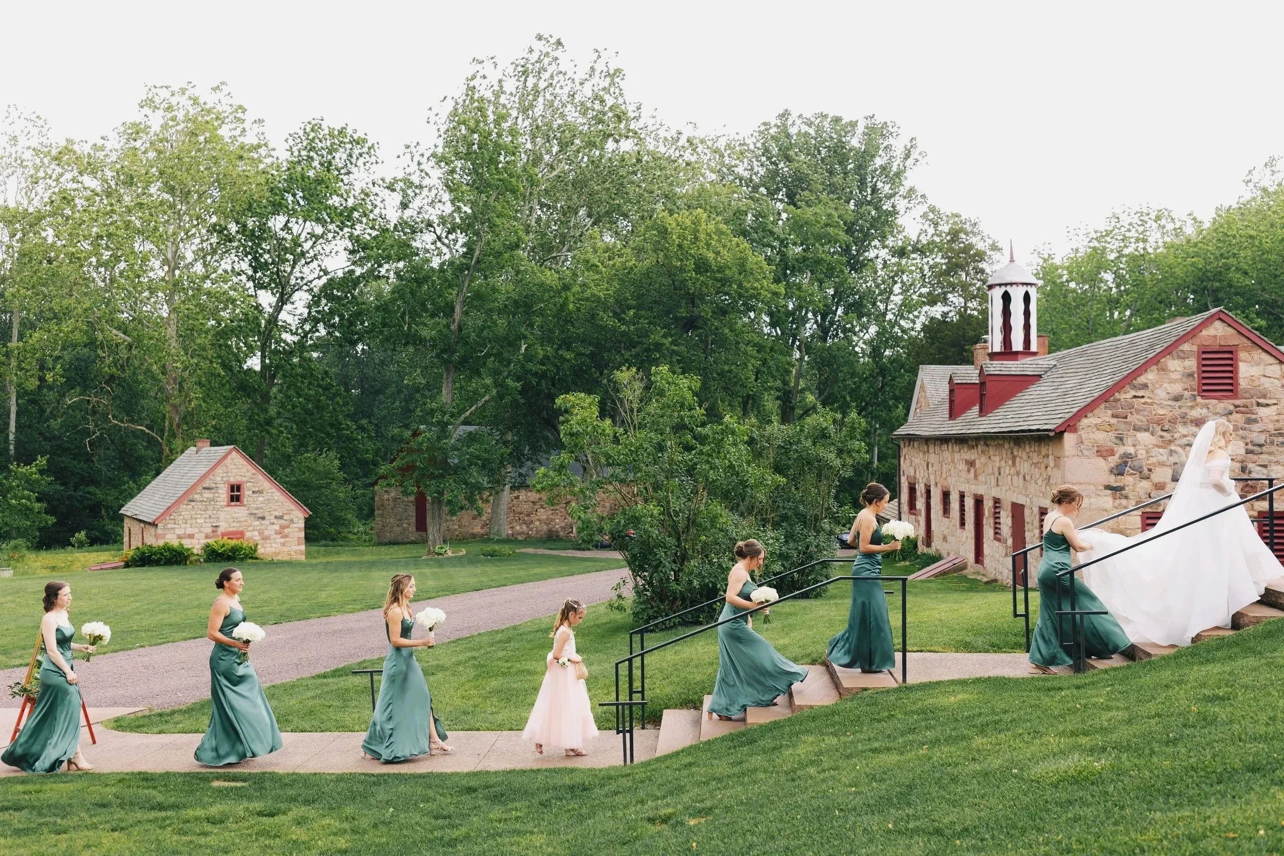 Bridesmaids and flower girl walking down the steps at an outdoor wedding ceremony on a grassy lawn, with stone barn buildings and tall green trees in the background.