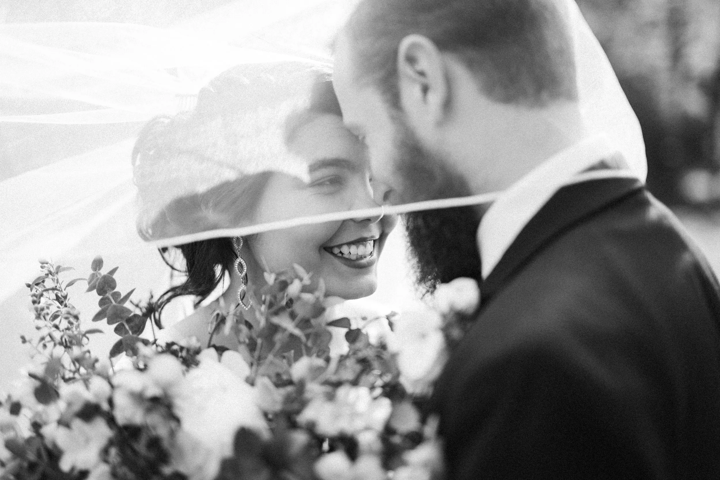 A black and white photo of a bride and groom smiling at each other, with the bride wearing earrings and a veil, holding a bouquet of flowers, and the groom wearing a suit, outdoors.