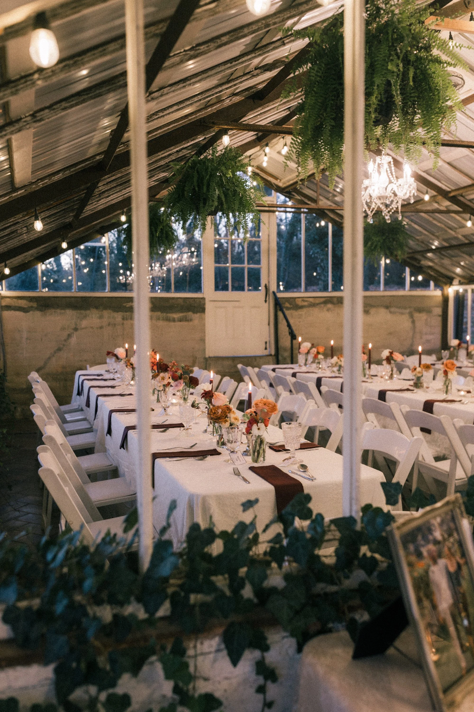 Indoor wedding or banquet reception with long tables draped in white tablecloths, decorated with floral arrangements and candles. Green hanging plants and crystal chandelier add elegant decor, with string lights and large windows visible in the background.