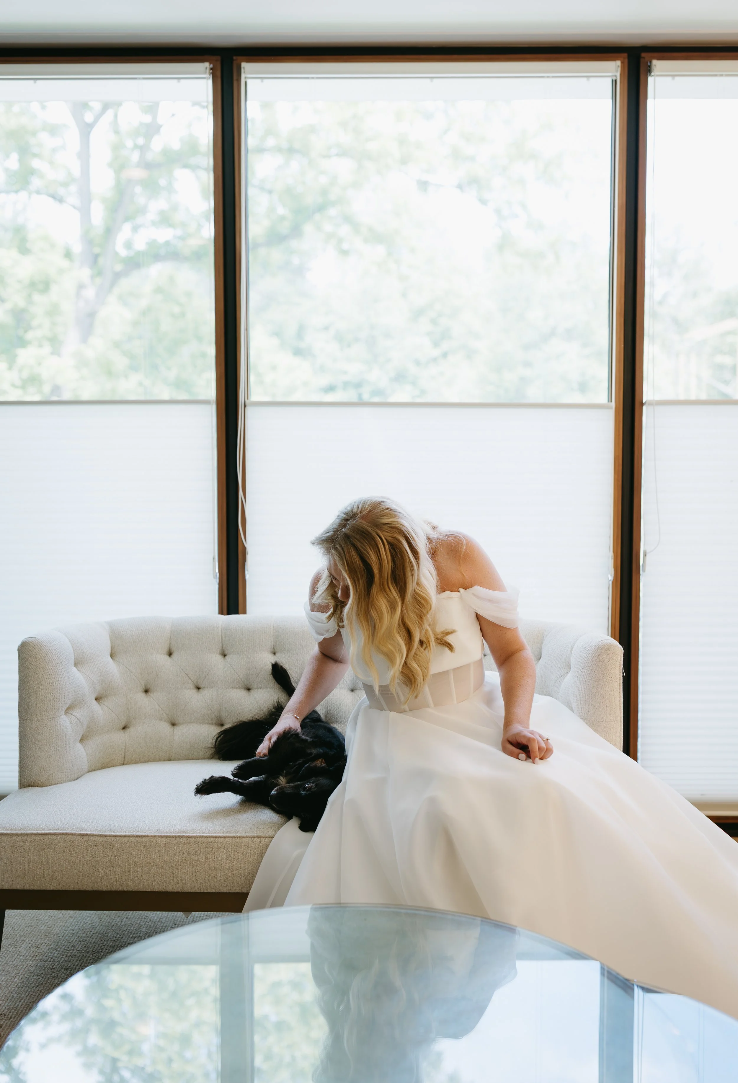 A woman in a white dress sitting on a beige sofa, petting a black dog lying on the sofa, with large windows and a view of trees outside in the background.