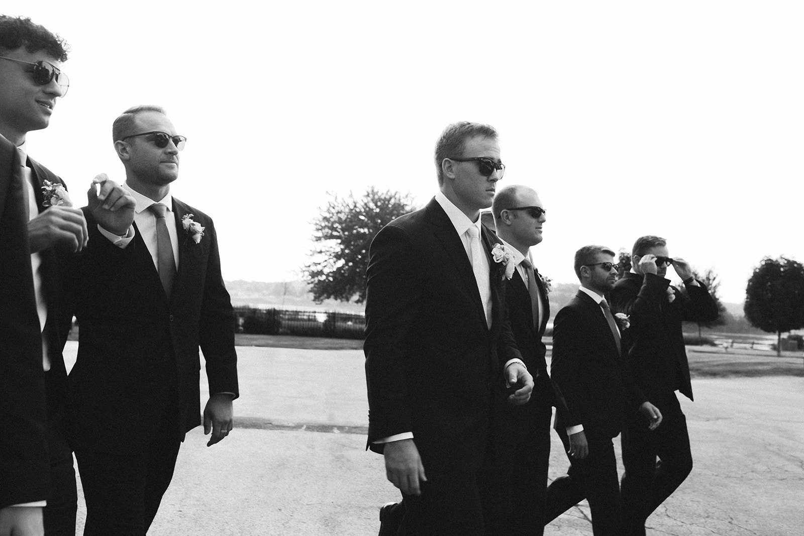 Six men in formal suits and sunglasses walking outdoors during daytime in a line.