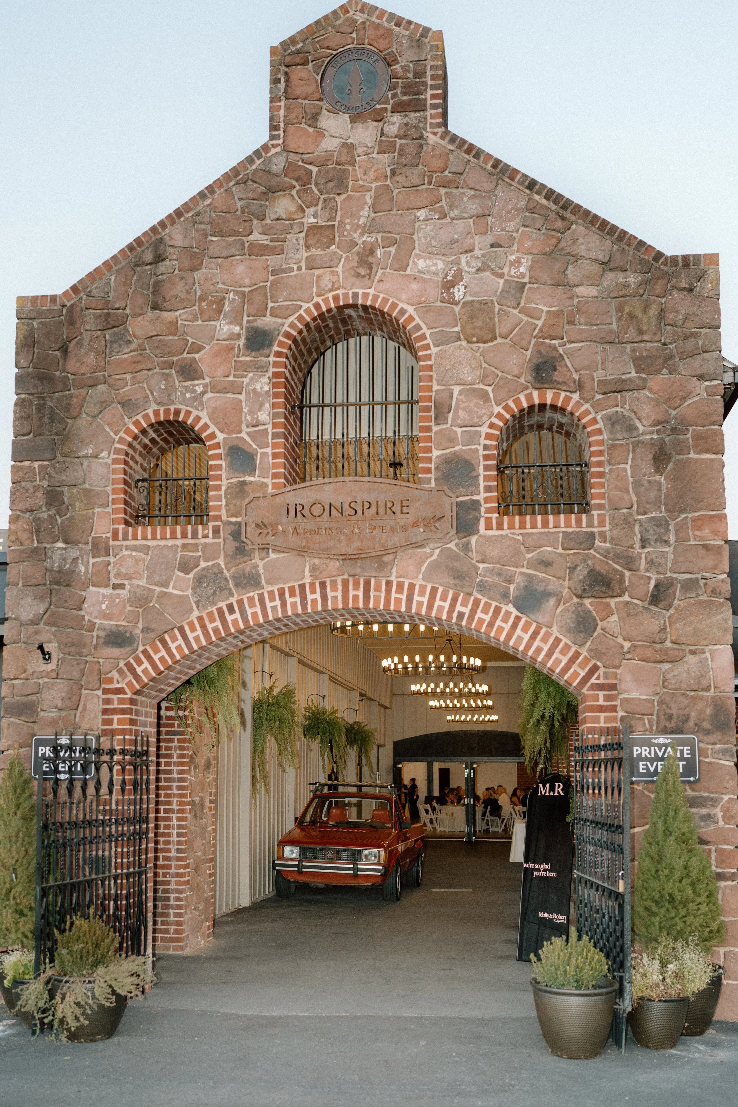 The entrance to a venue with a stone brick archway reading 'IRONSPIRE WEDDING & EVENTS,' leading inside to a decorated interior with chandeliers and greenery, with a red vintage car parked in front and signs indicating private event areas.