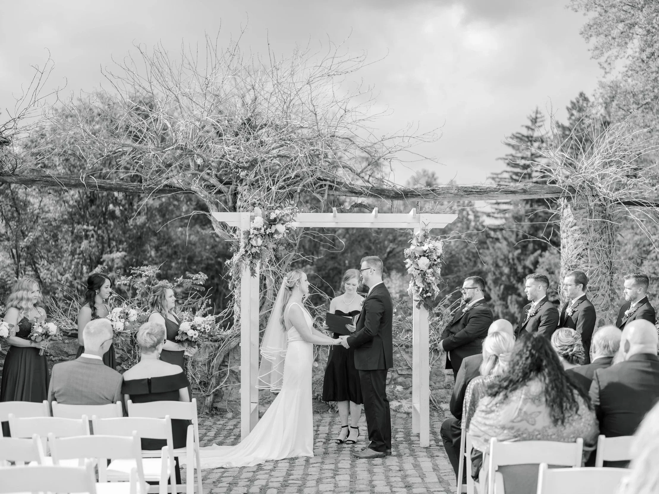 Black and white photo of an outdoor wedding ceremony with a bride and groom exchanging vows under a wooden arch decorated with flowers, surrounded by wedding guests.