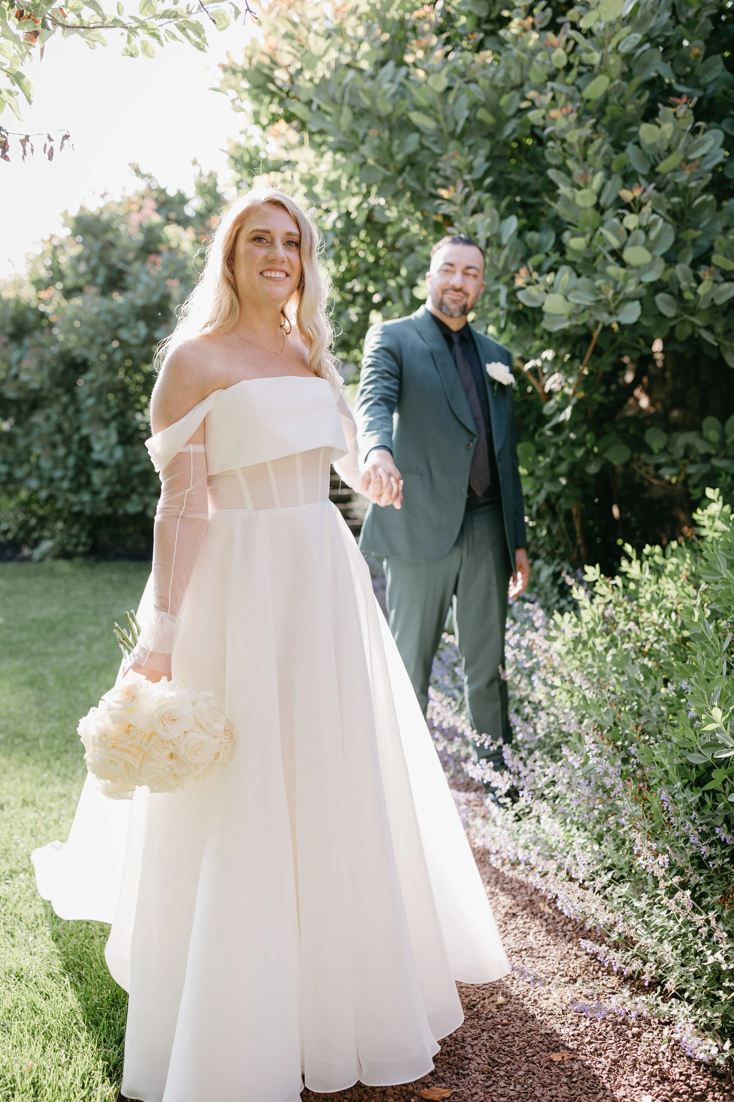 A smiling woman in a white off-the-shoulder wedding dress holding a bouquet of white roses in a garden, with a man in a teal suit and black shirt in the background, surrounded by greenery and flowering plants.