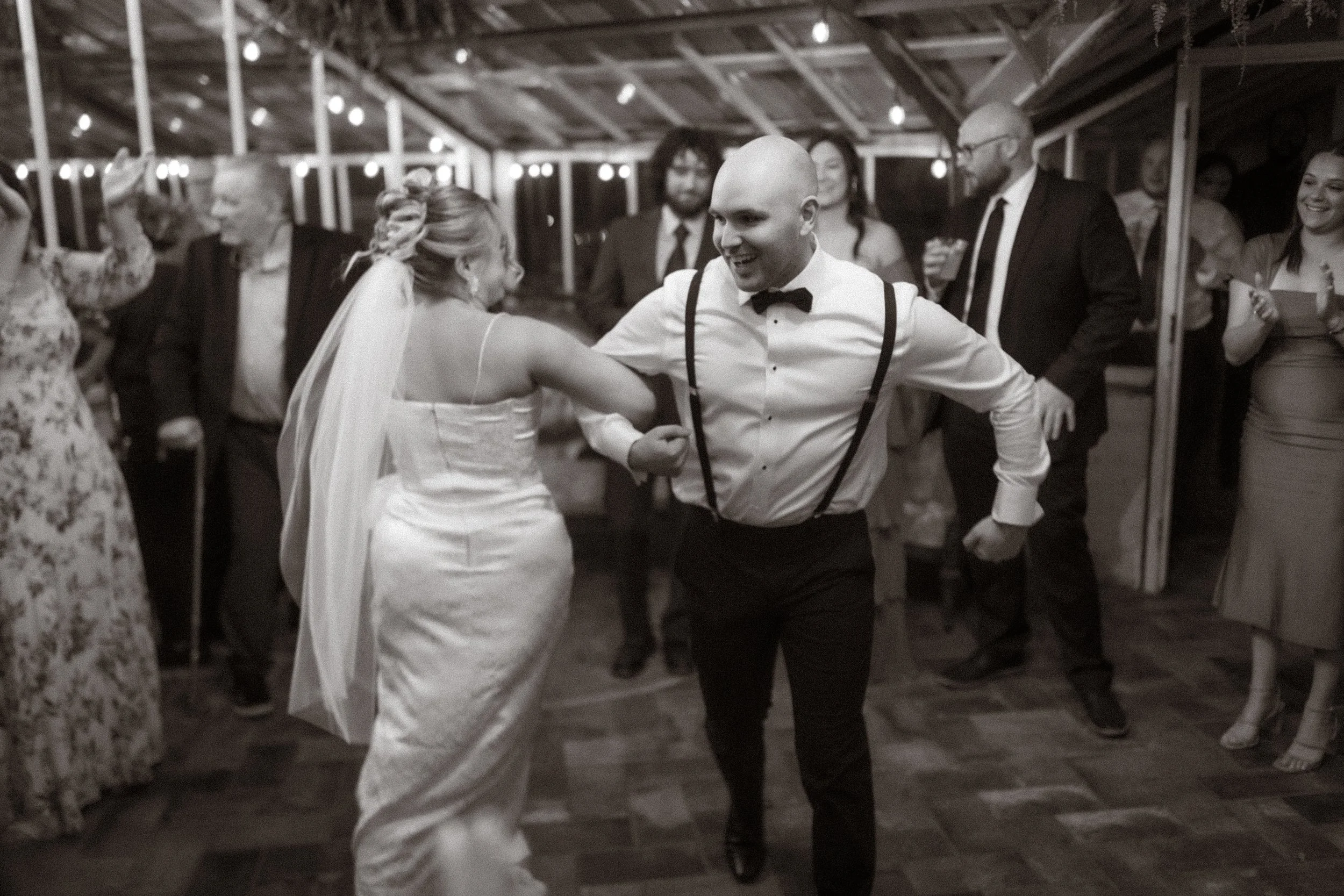 A group of people dancing and celebrating at a wedding reception or party in a barn or rustic venue with string lights overhead.