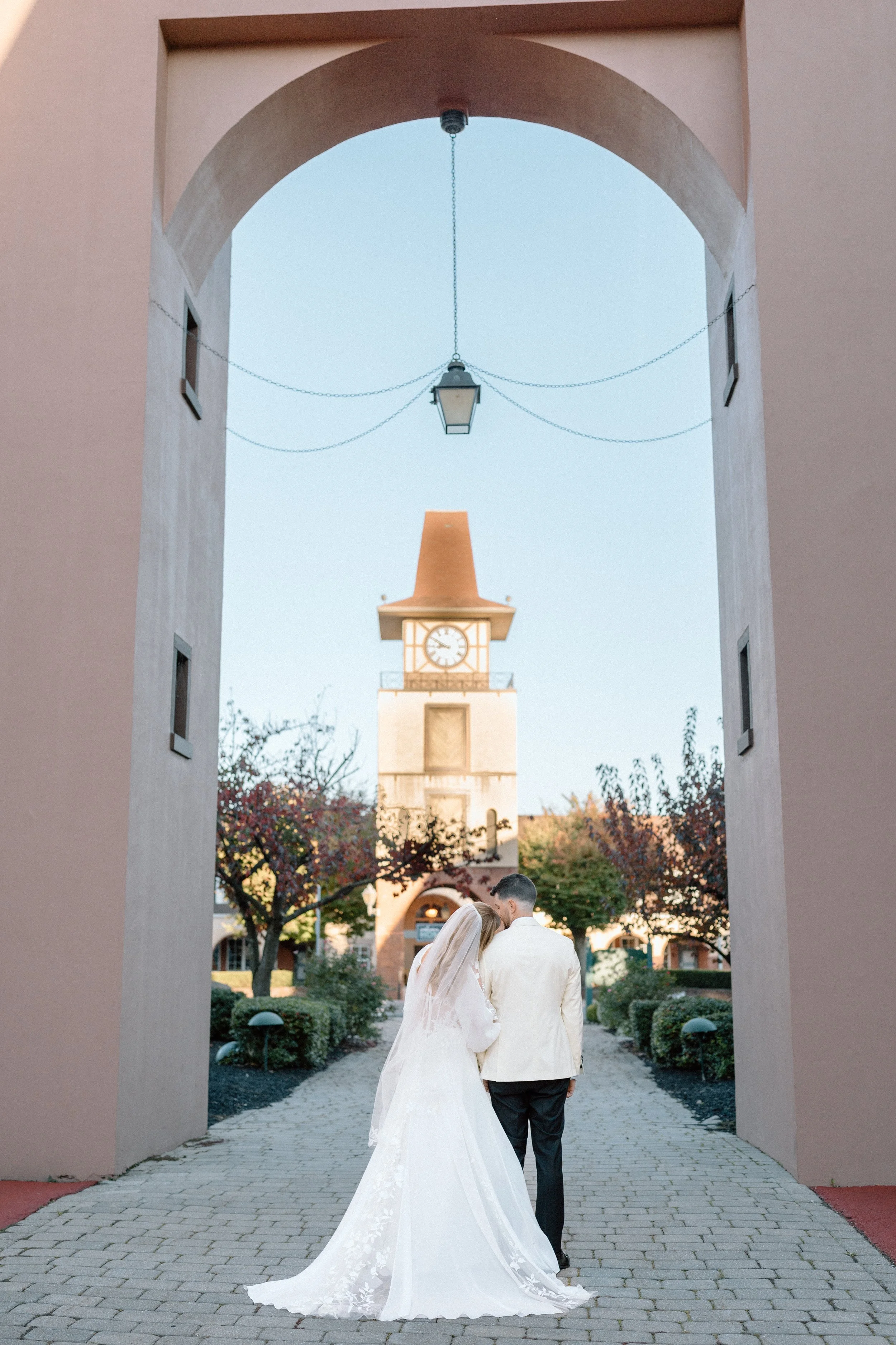 A bride and groom walking together under an archway, with a clock tower and autumn trees in the background.