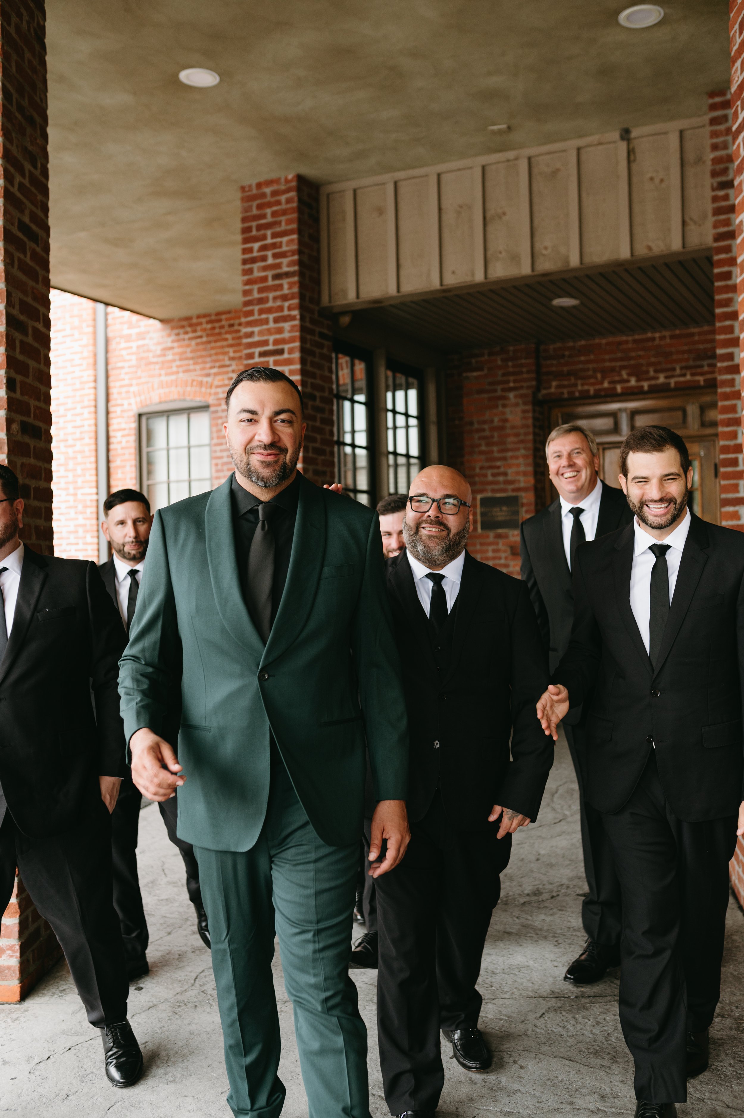A group of men dressed in tuxedos walking together in an industrial-style brick building with large windows and concrete floors. The man at the front is wearing a green suit and smiling.